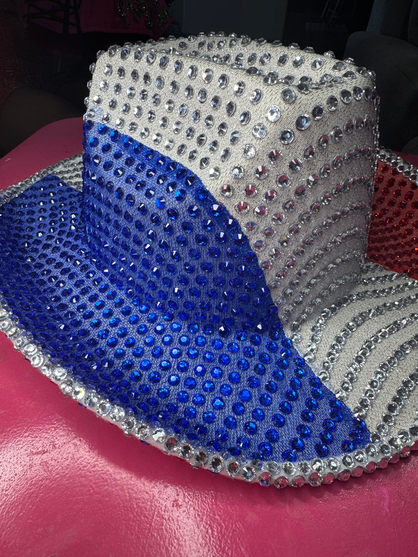 The Viva La Bombshell Burlesque Rhinestoned Cowboy Hat features silver, blue, and red rhinestones in vibrant sections, creating a patriotic look. It’s pictured resting on a shiny pink surface.