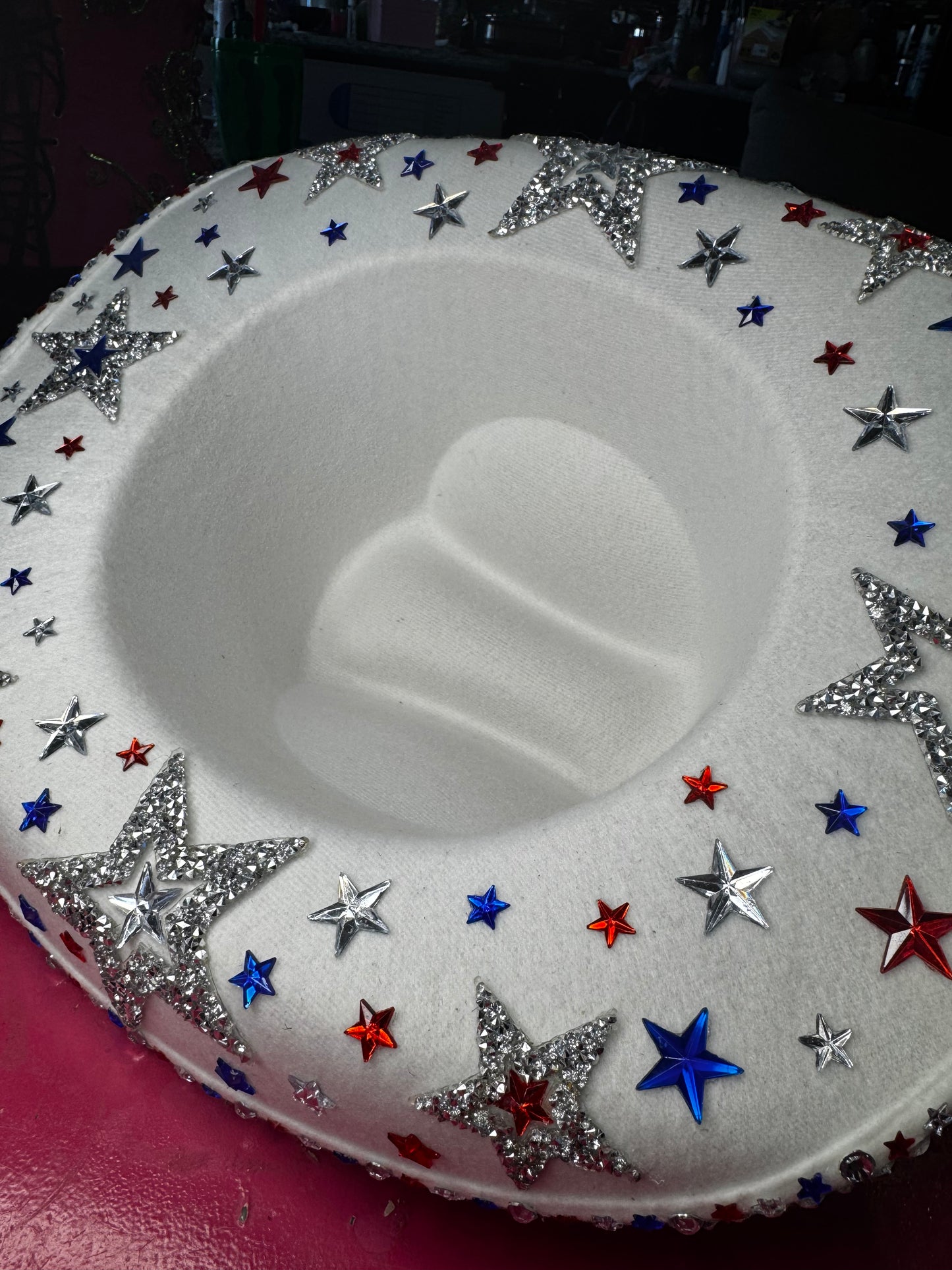 The Viva La Bombshell Burlesque Rhinestoned Cowboy Hat is a white, plush seat with an indented center, adorned with silver, red, and blue star designs for a patriotic, festive look on a pink surface.