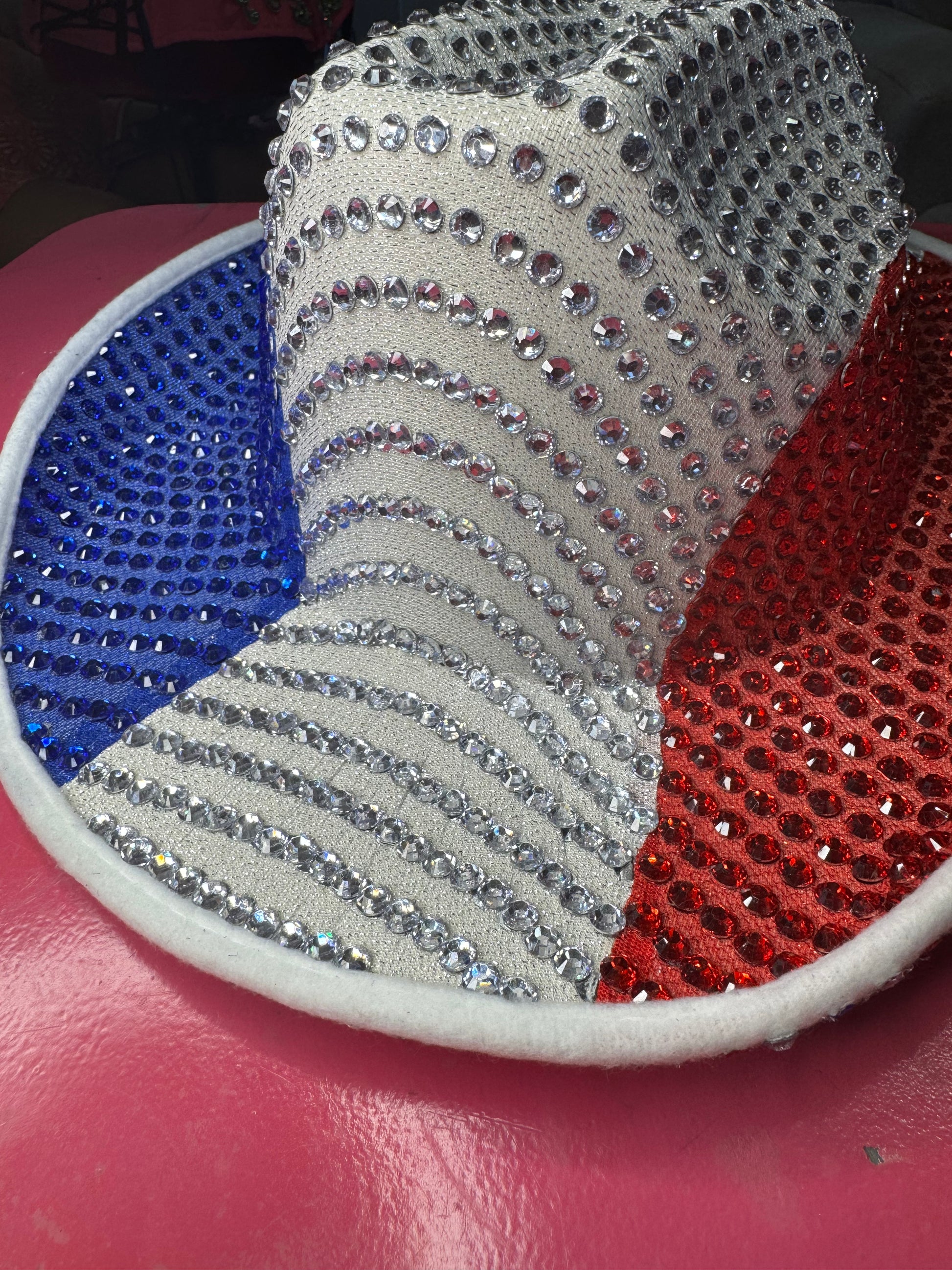 The Viva La Bombshell Burlesque Rhinestoned Cowboy Hat, adorned with blue, white, and red sparkling rhinestones, rests on a shiny pink surface.