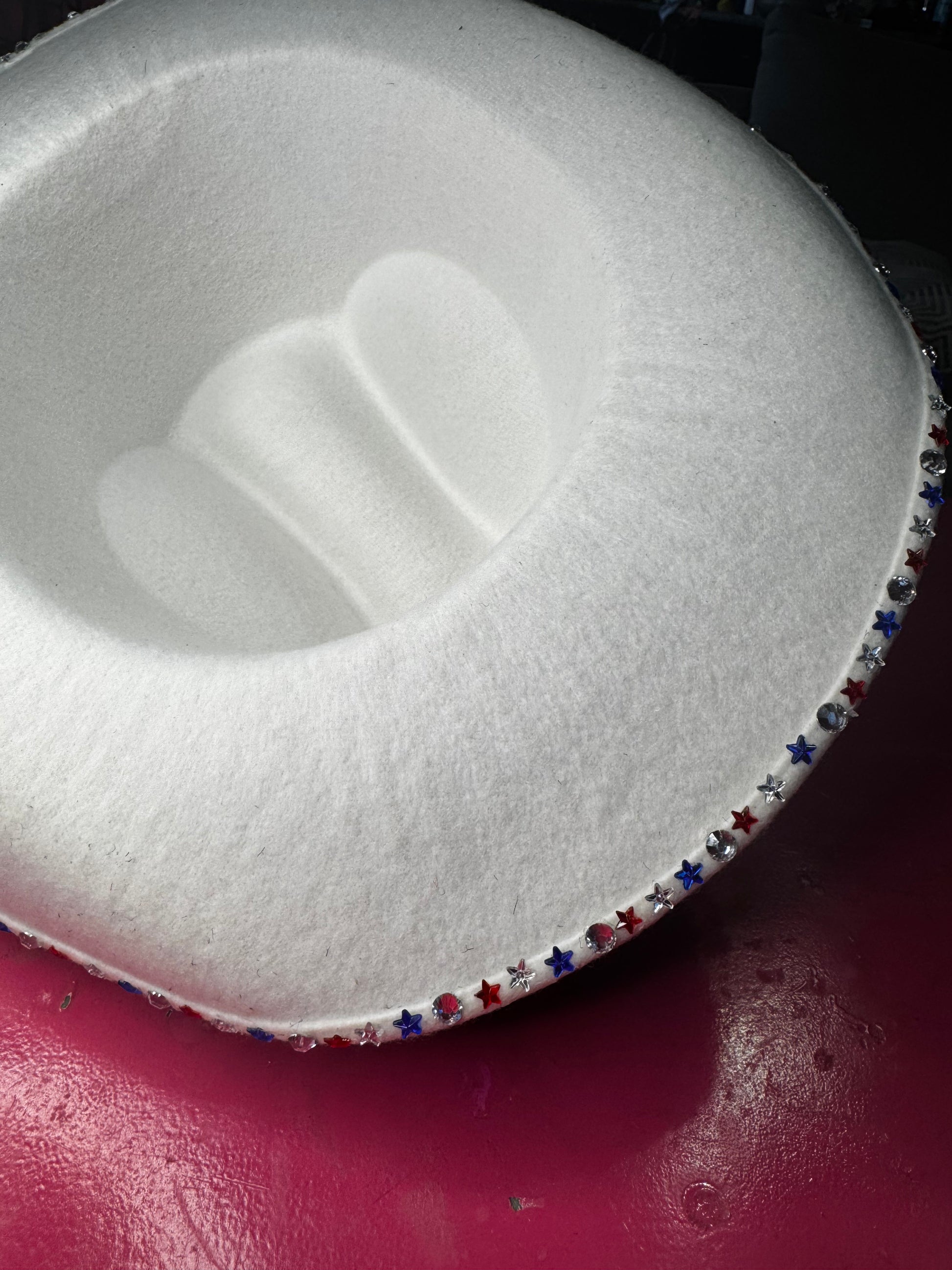 A white, plush, rounded chair rests on a glossy pink floor. Red, blue, and silver star accents along its edge evoke the sparkle of Viva La Bombshell Burlesque’s Rhinestoned Cowboy Hat.