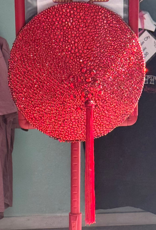 The Titan Tip Tit by Viva La Bombshell Burlesque: a large, round hand fan adorned with red rhinestones and a central red tassel, reminiscent of a giant pastie, displayed upright against a muted green and black background.