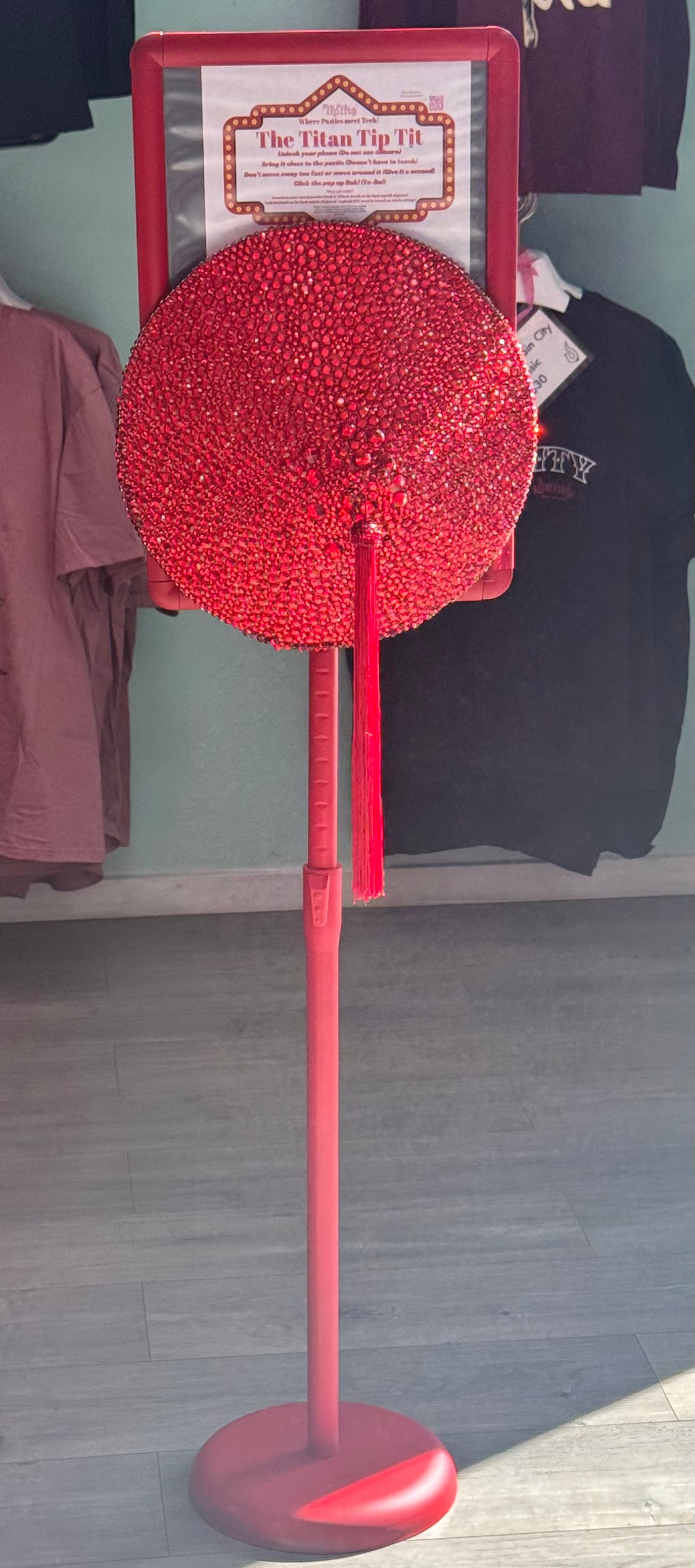 The Titan Tip Tit by Viva La Bombshell Burlesque is a red, circular rhinestone-covered tip jar with a tassel on a red stand. An inviting sign tops it, while festival merch shirts hang in the background.