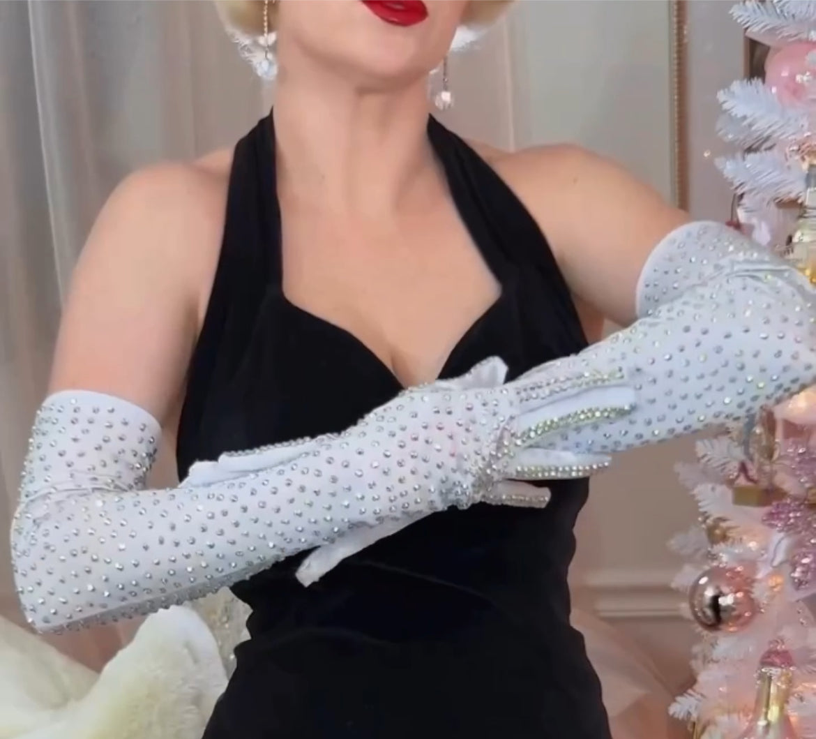 Wearing Viva La Bombshell Burlesque’s Classic Rhinestone Gloves, a woman in a black halter dress and red lipstick adds a touch of glamour as she stands beside a white Christmas tree.