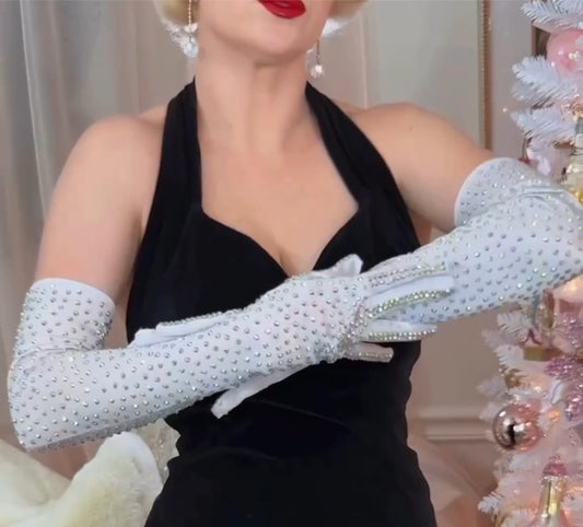 Wearing Viva La Bombshell Burlesque’s Classic Rhinestone Gloves, a woman in a black halter dress and red lipstick adds a touch of glamour as she stands beside a white Christmas tree.