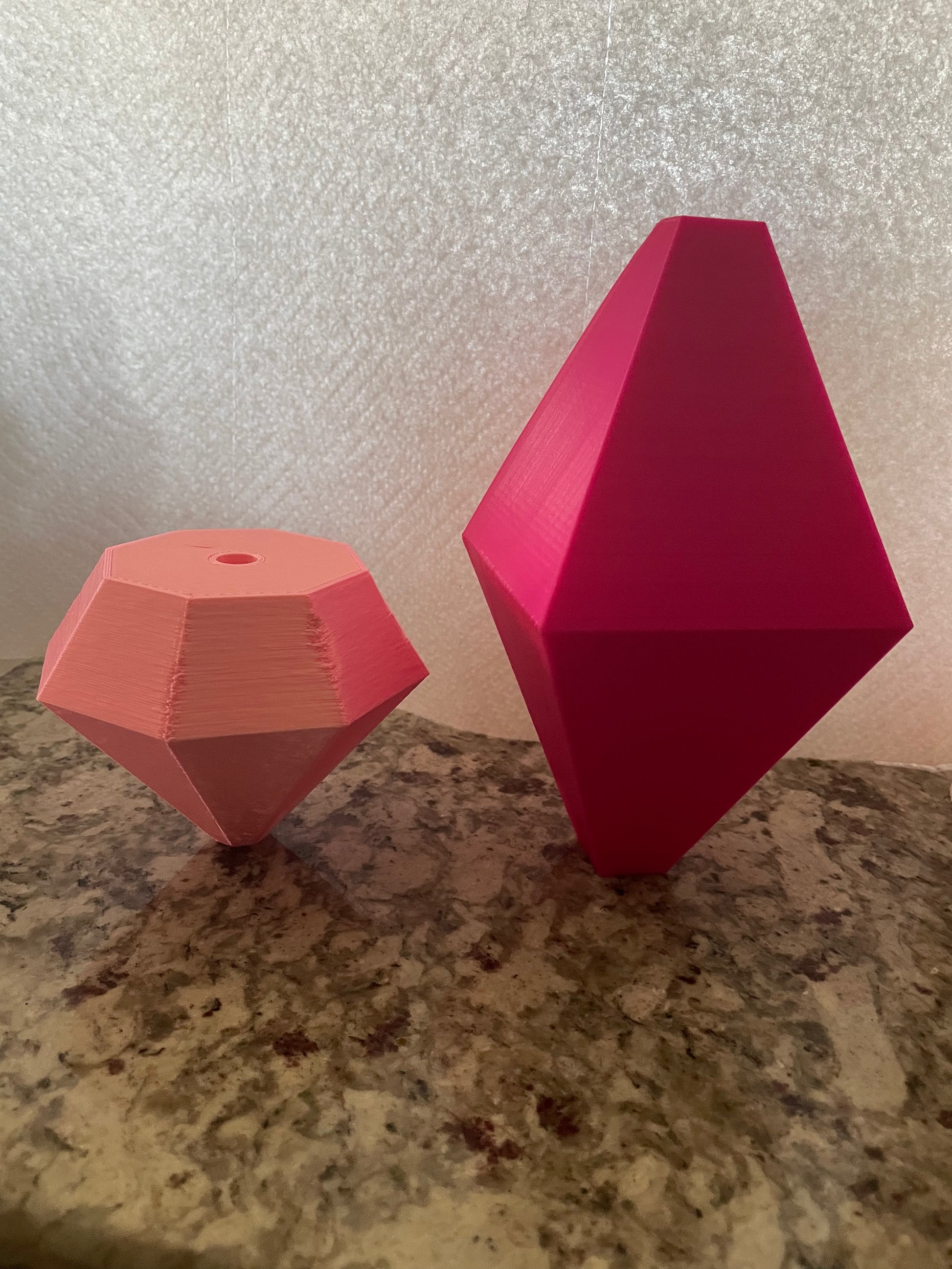 Two geometric 3D printed charms from Viva La Bombshell Burlesque’s Boa Charms - Boa Attachments sit on granite: a small, light pink faceted charm with a top hole and a larger, bright pink diamond shape against a textured background.