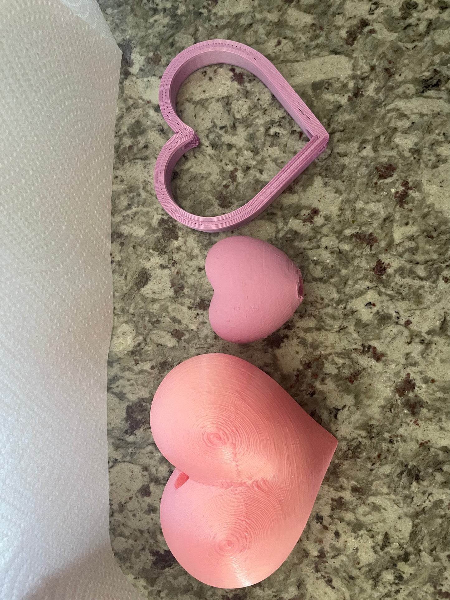 Three pink 3D printed Boa Charms - Boa Attachments by Viva La Bombshell Burlesque, each with unique heart-shaped designs and sizes, are displayed on a speckled countertop beside a sheet of white paper towel.