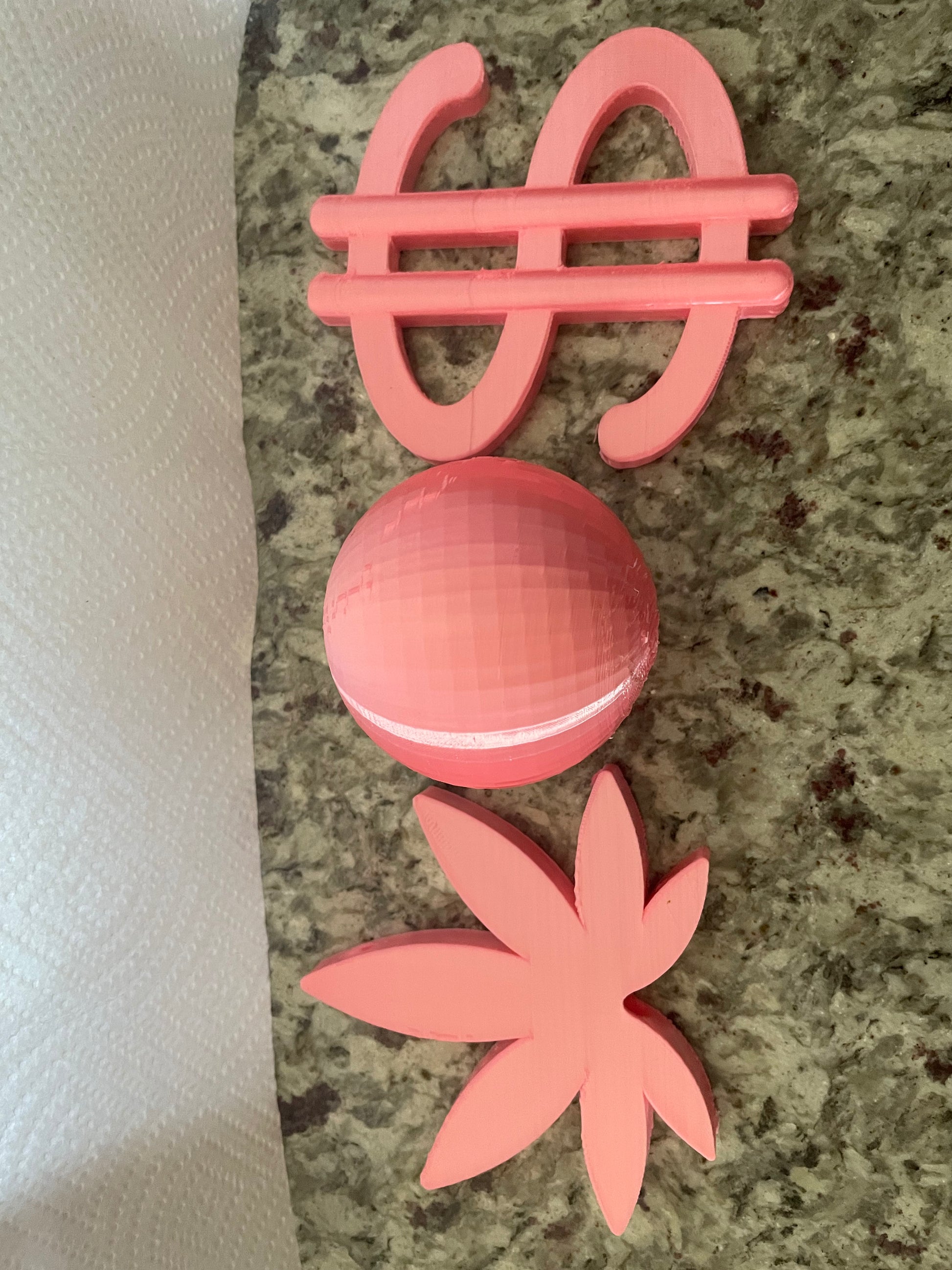 Three pink 3D-printed Viva La Bombshell Burlesque Boa Charms—dollar sign, faceted oval, and stylized cannabis leaf—sit on a countertop, showcasing unique Boa Attachments.