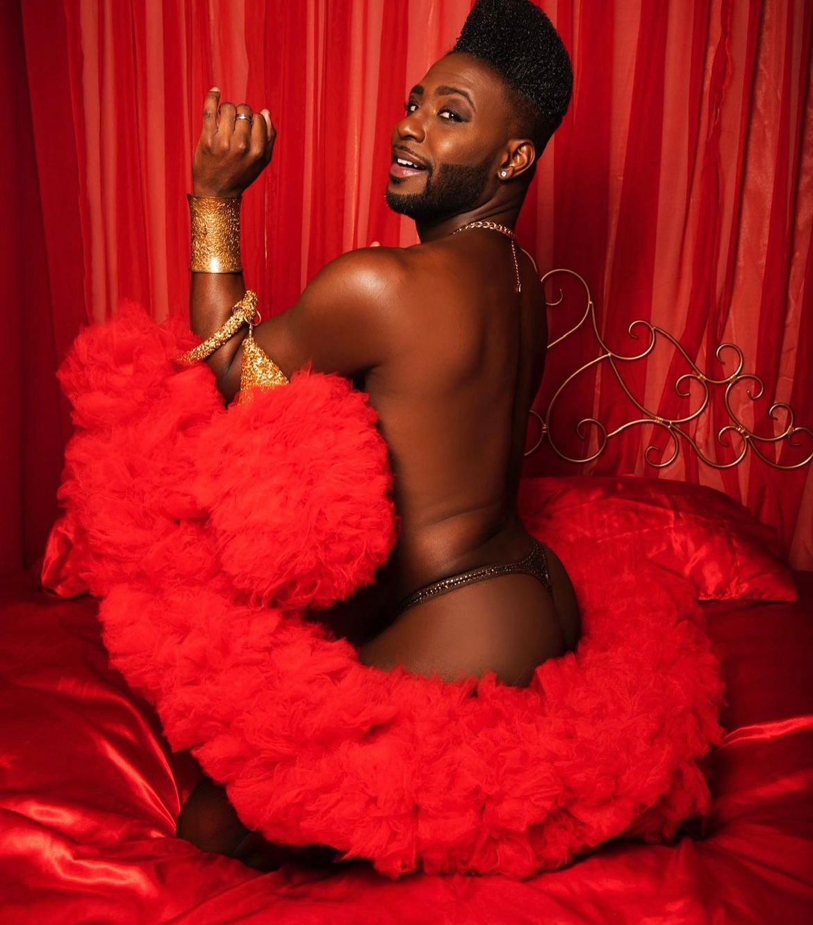 A confident model poses on a red bed, draped in Viva La Bombshell Burlesque's The De'Vil Tulle Boa, with gold accessories and bold makeup, against rich red curtains.