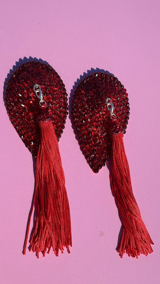 Viva La Bombshell Burlesque's Teardrop Pasties feature red rhinestones, long red tassels, and a silver clasp. Reusable and glamorous, they're perfect for performances or special occasions. Shown on a pink background.