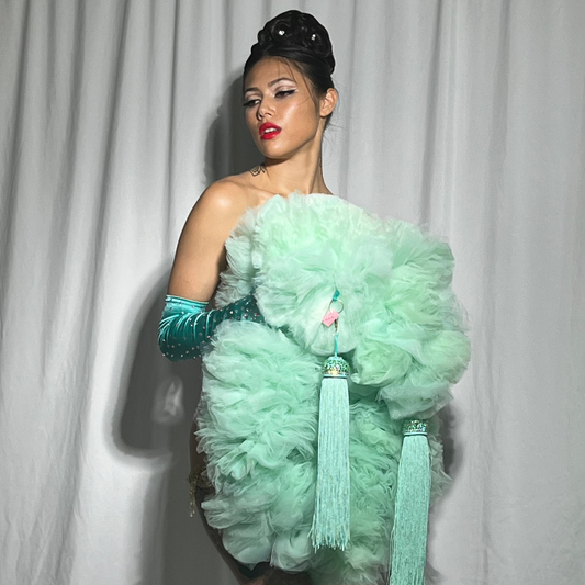 A person with styled hair and bold red lipstick poses in front of white curtains, wearing bright turquoise gloves and Viva La Bombshell Burlesque's "The Dazzle - Tulle Boa," a large, fluffy mint-green costume adorned with tassels.