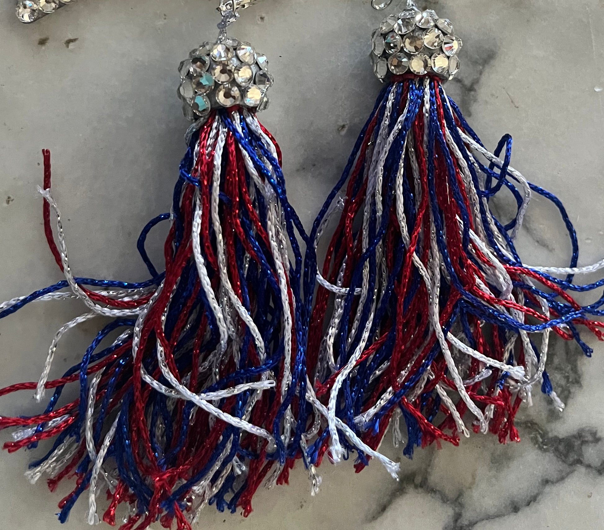 The Viva La Bombshell Burlesque Pastie Tassels feature shiny rhinestone toppers with metallic red, blue, and silver thread strands, displayed on a light gray marble surface.