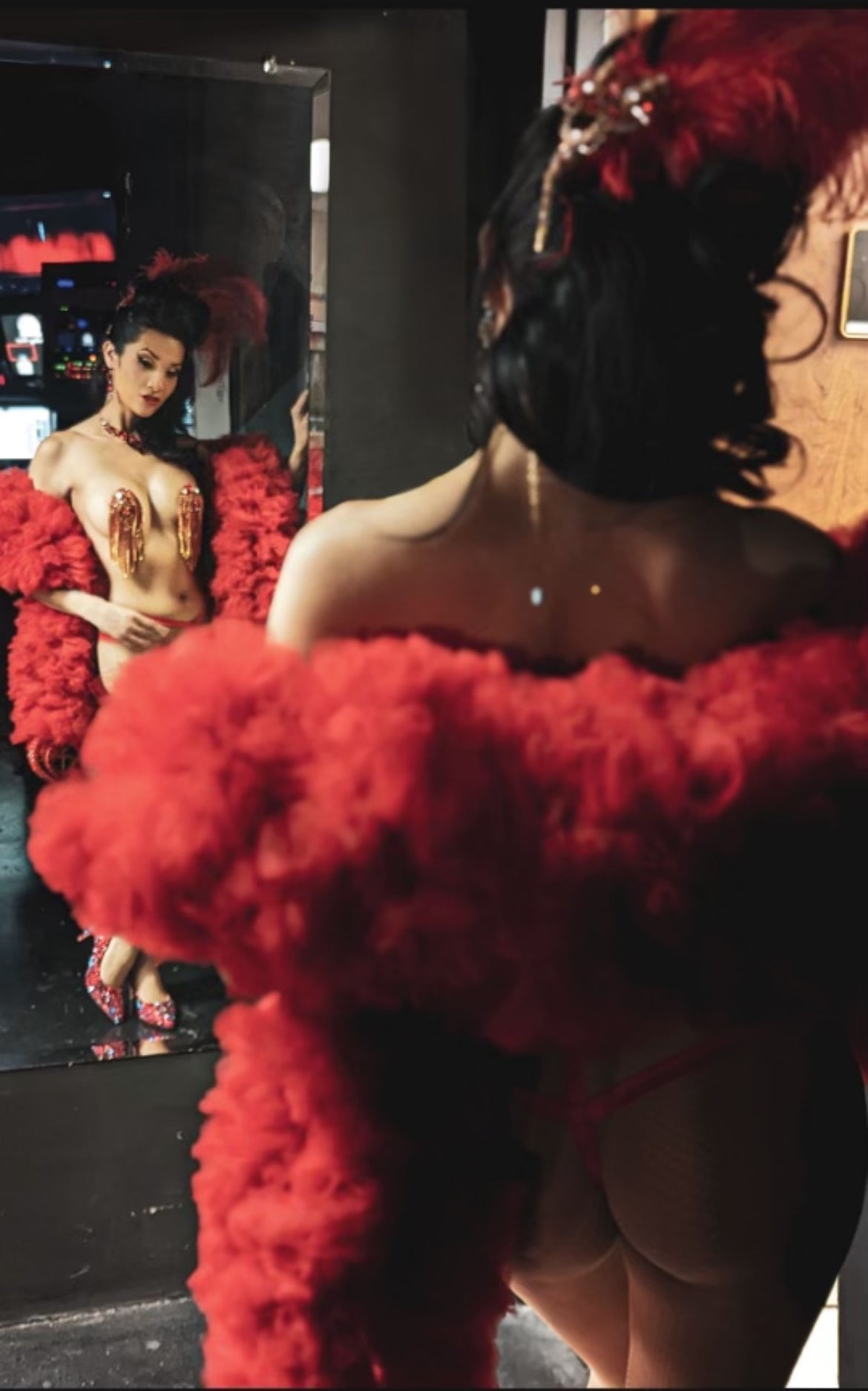 A person in a red feathered costume and headpiece admires their reflection, back to the camera, showcasing sparkling accessories and The Devine - Tulle Boa by Viva La Bombshell Burlesque.