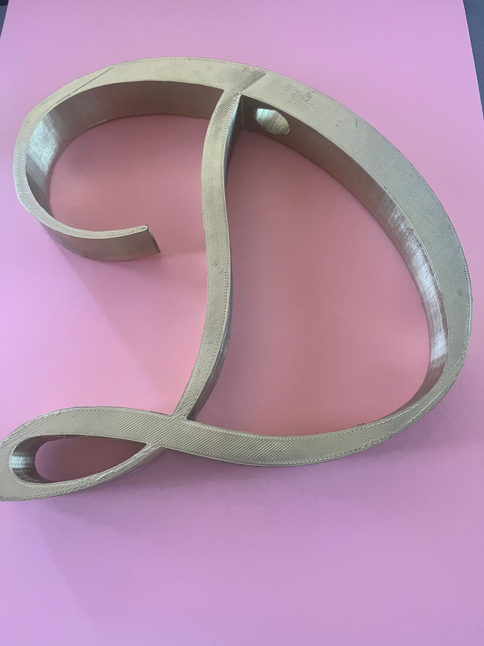 A large, gold 3D letter D from the Letter Charms - Boa Attachments by Viva La Bombshell Burlesque sits on a flat pink surface, appearing lightweight and possibly made of plastic or 3D-printed material.