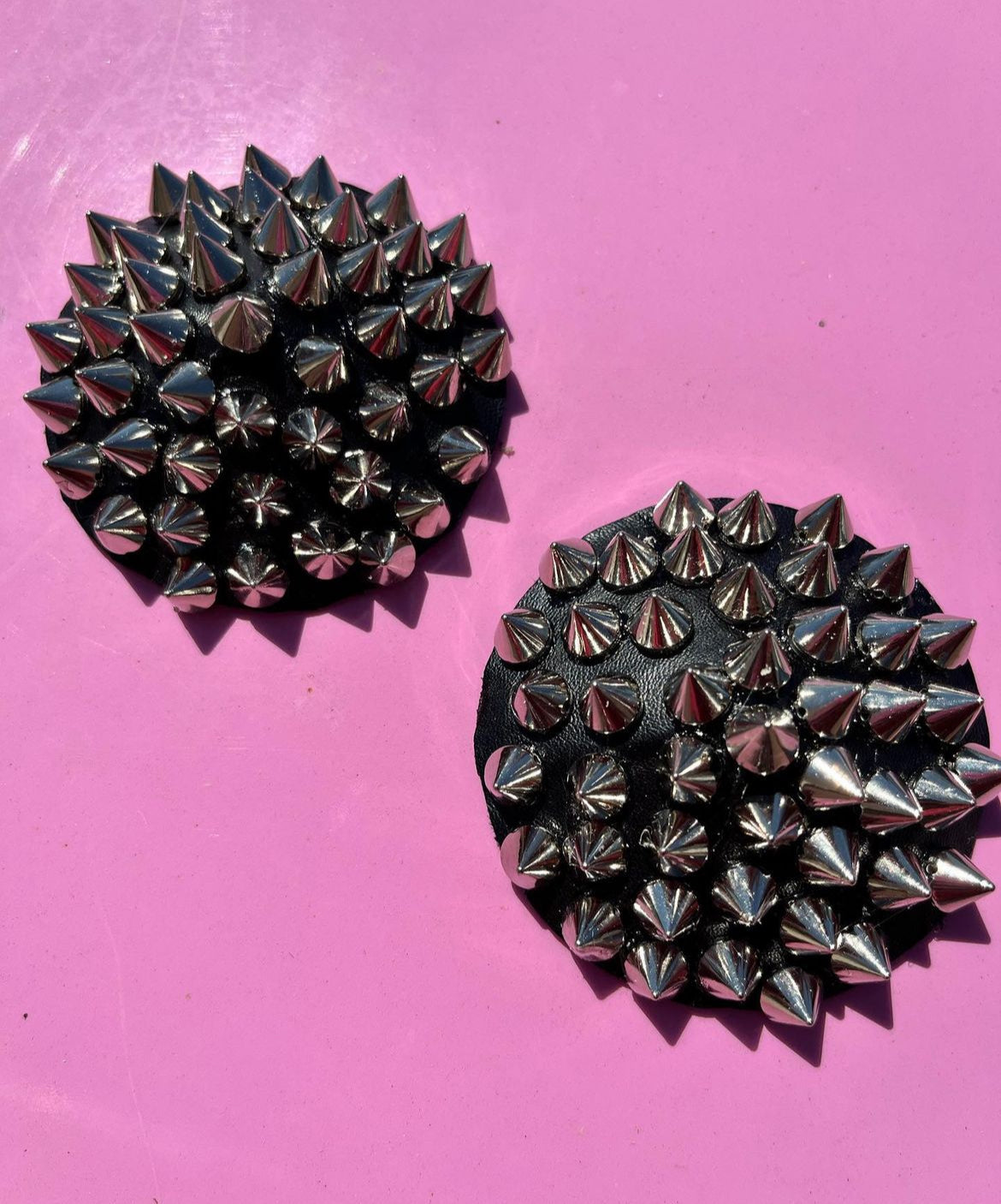 Viva La Bombshell Burlesque Spiked Pasties, featuring round shapes covered in shiny silver spikes, rest on a bright pink surface and cast small shadows.