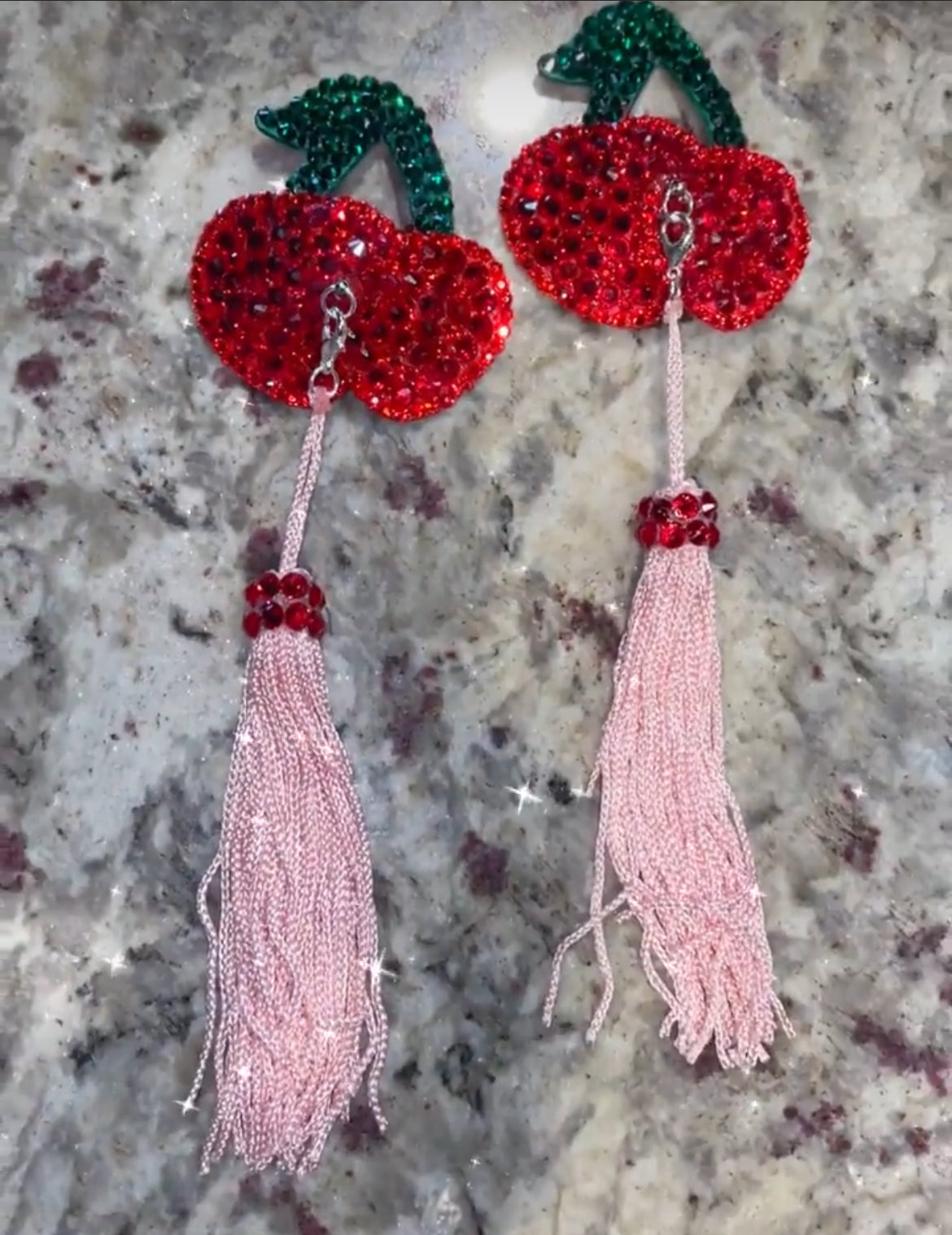 Cherry Pasties by Viva La Bombshell Burlesque feature red sequins, green leaves, and long pink beaded tassels, capturing a playful style and shown beautifully on a marble surface.