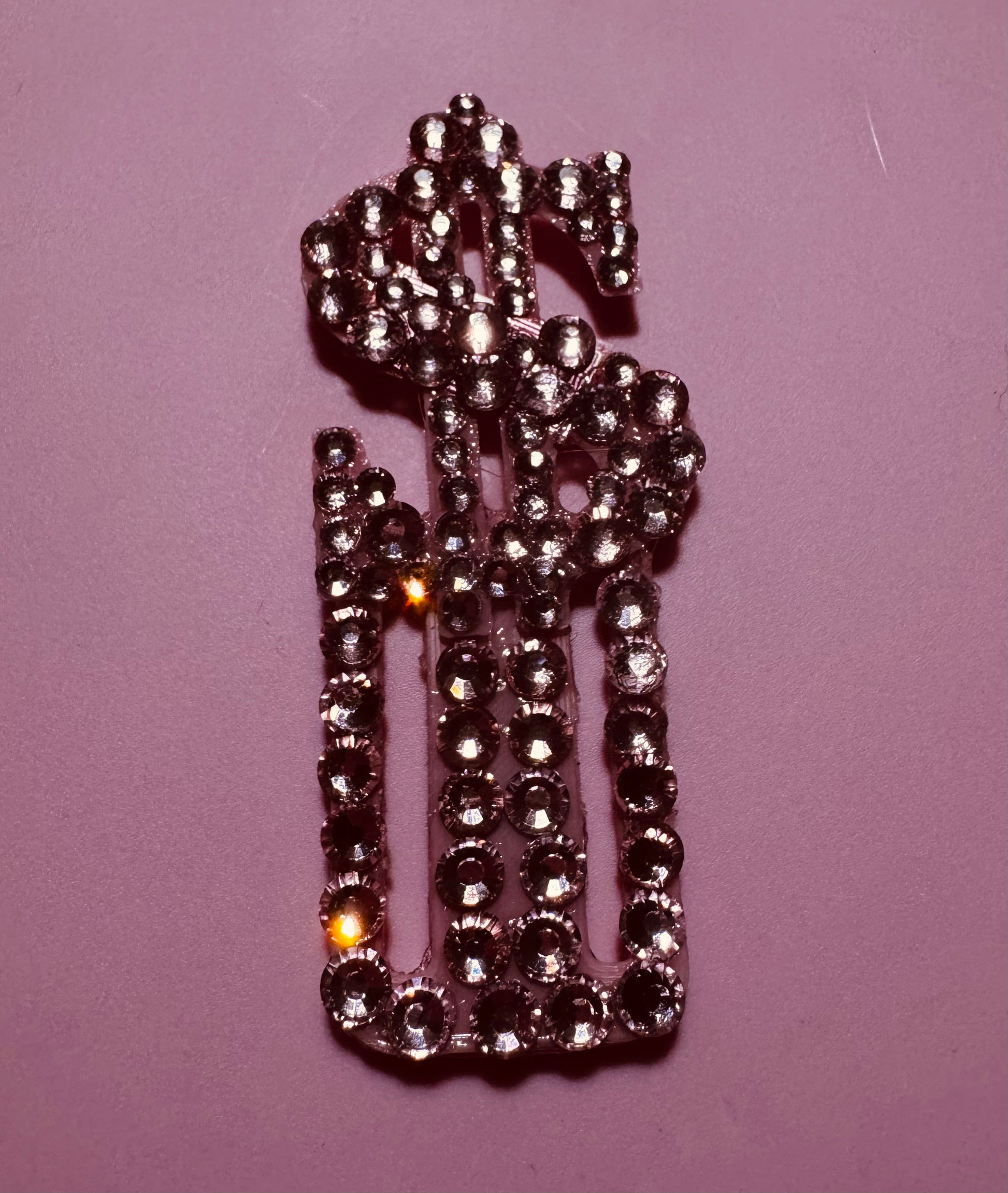 A rhinestone dollar sign brooch from Viva La Bombshell Burlesque is displayed on pink, echoing wearable tech trends like TipSlider for digital tipping, with light dancing across the stones.