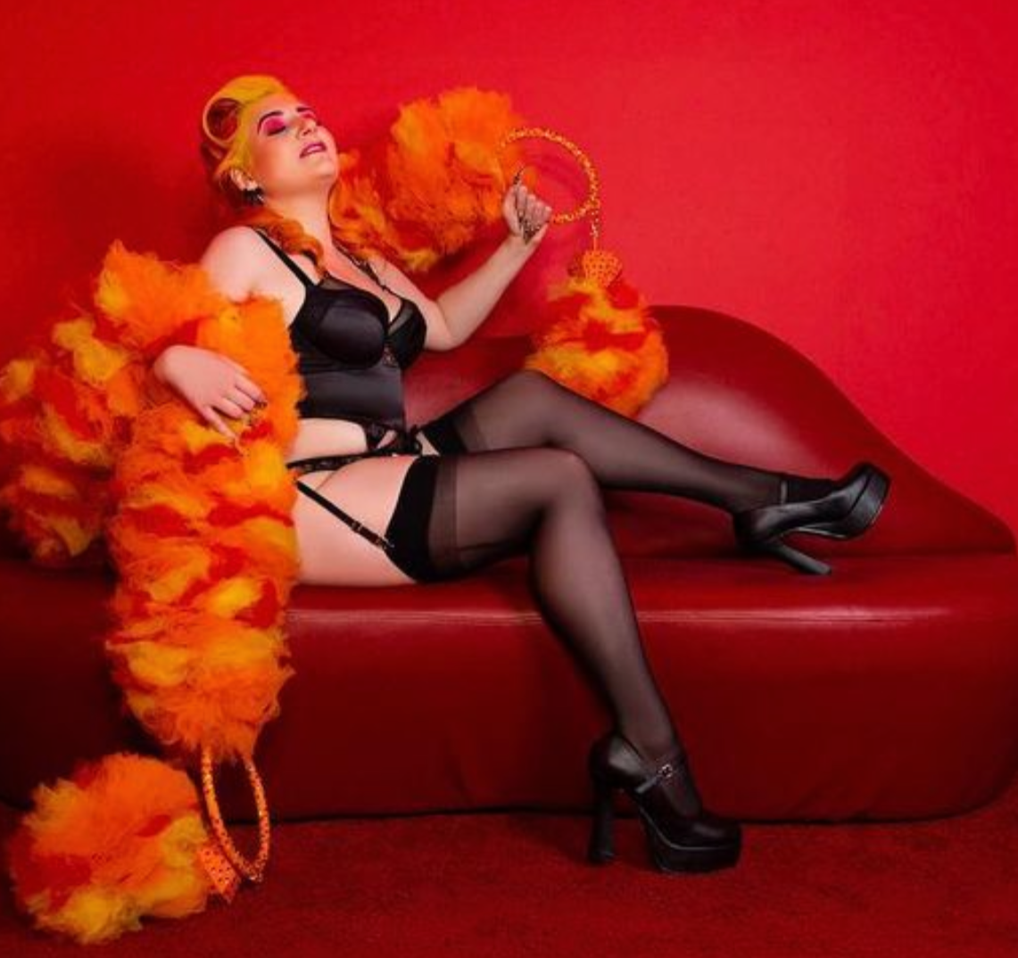 Wearing black lingerie and stockings, a person lounges on a red chaise with orange curlers, holding Viva La Bombshell Burlesque's The De'Vil - Tulle Boa and vibrant feather fans, set against a bold red background.