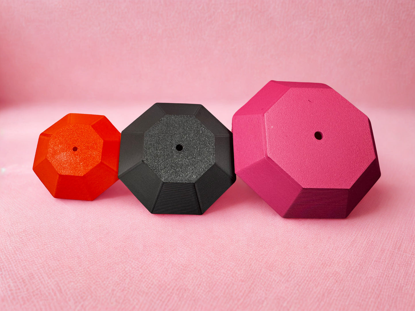 Three Tassel Toppers - Boa by Viva La Bombshell Burlesque, each with a central hole, are arranged from smallest to largest in red, black, and pink against a soft pink background—ideal for DIY 3D printed rhinestone tassel toppers.