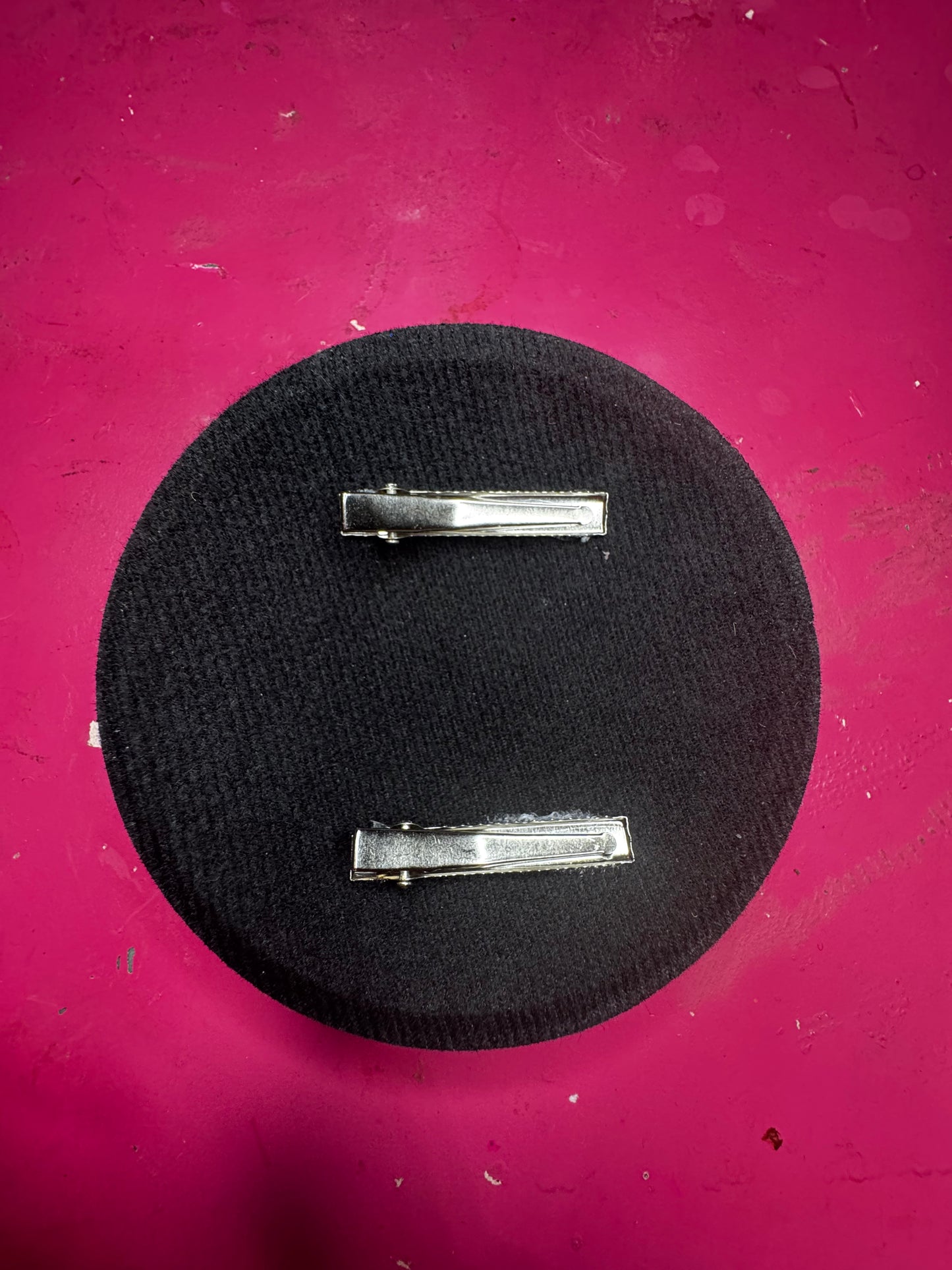 The Viva La Bombshell Burlesque Tufted Heart Fascinator, a handmade black fabric hair accessory with two metal clips, sits atop a bright pink textured surface, offering chic style and everyday practicality.