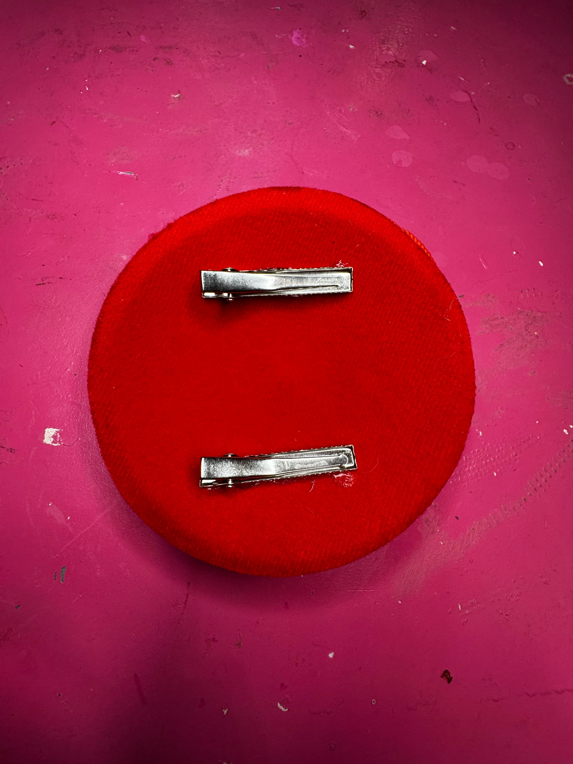 The Tufted Heart Fascinator by Viva La Bombshell Burlesque, a round red fabric hair accessory with two silver clips, rests on a pink, slightly stained surface.