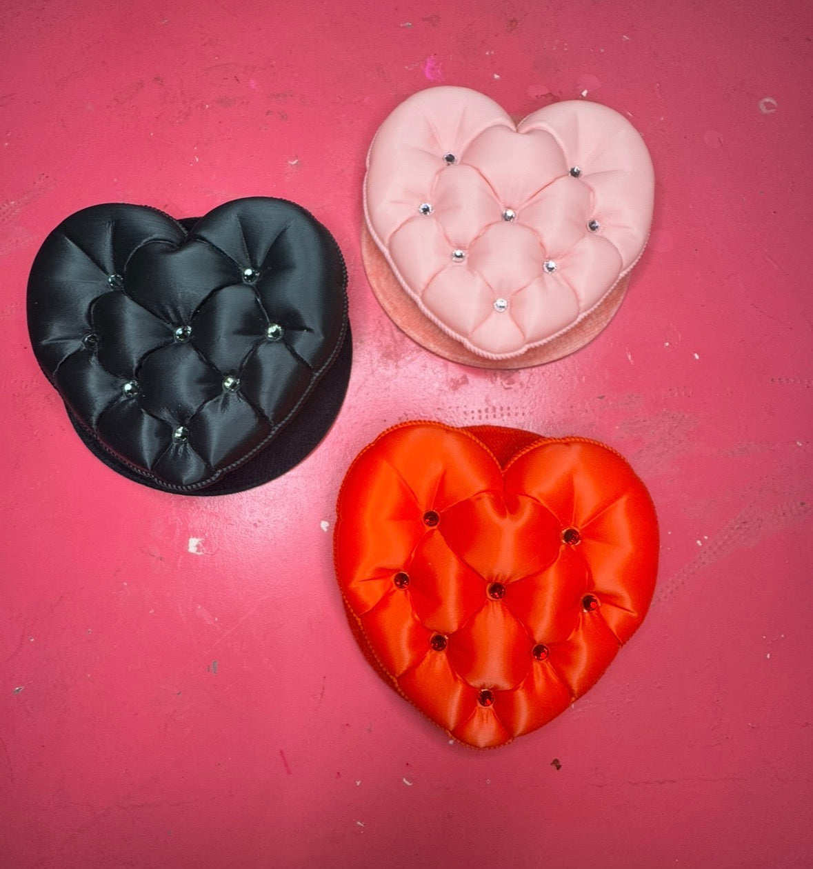 Three heart-shaped, tufted cushions with button details in black, pink, and red from Viva La Bombshell Burlesque's Tufted Heart Fascinator line are arranged on a pink surface, adding flair to any handmade hair accessory collection.