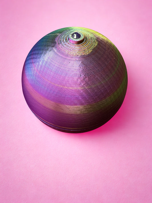 A vibrant, round metallic spinning top with iridescent purple, green, and gold hues sits on a bright pink surface next to the Viva La Bombshell Burlesque Boobie Box custom pastie storage box.