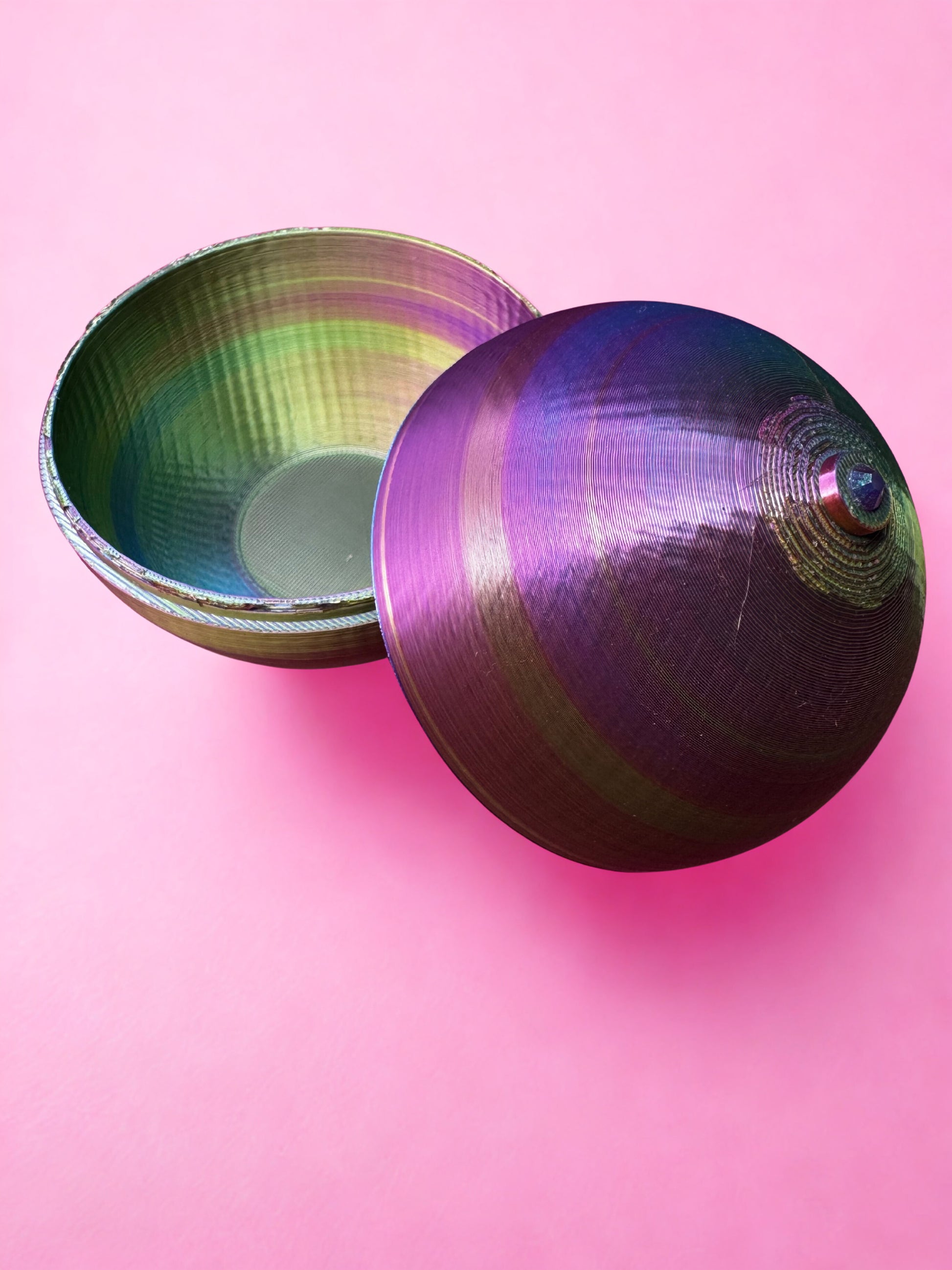The Boobie Box by Viva La Bombshell Burlesque, a round iridescent container shimmering with green, purple, and blue hues, sits on a pink surface with its lid slightly open to reveal its empty interior.