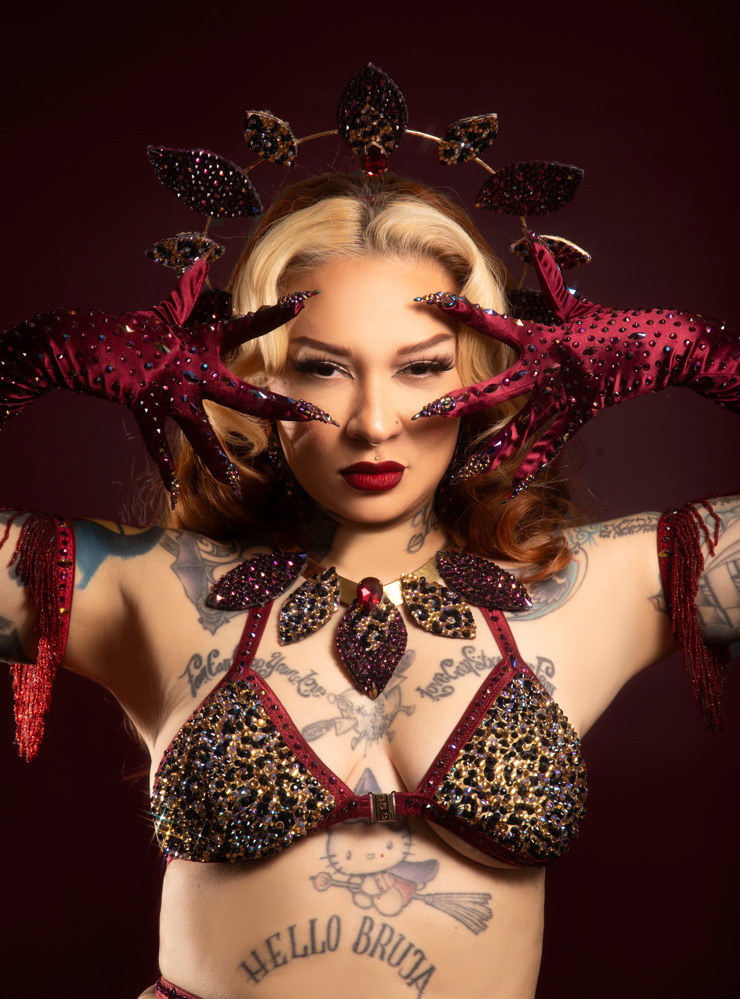A tattooed woman in an ornate, jeweled burgundy bra and gloves poses with her hands framing her face. She wears a matching headpiece, bold makeup, and has a Hello Bruta tattoo featuring Hello Kitty on her chest.