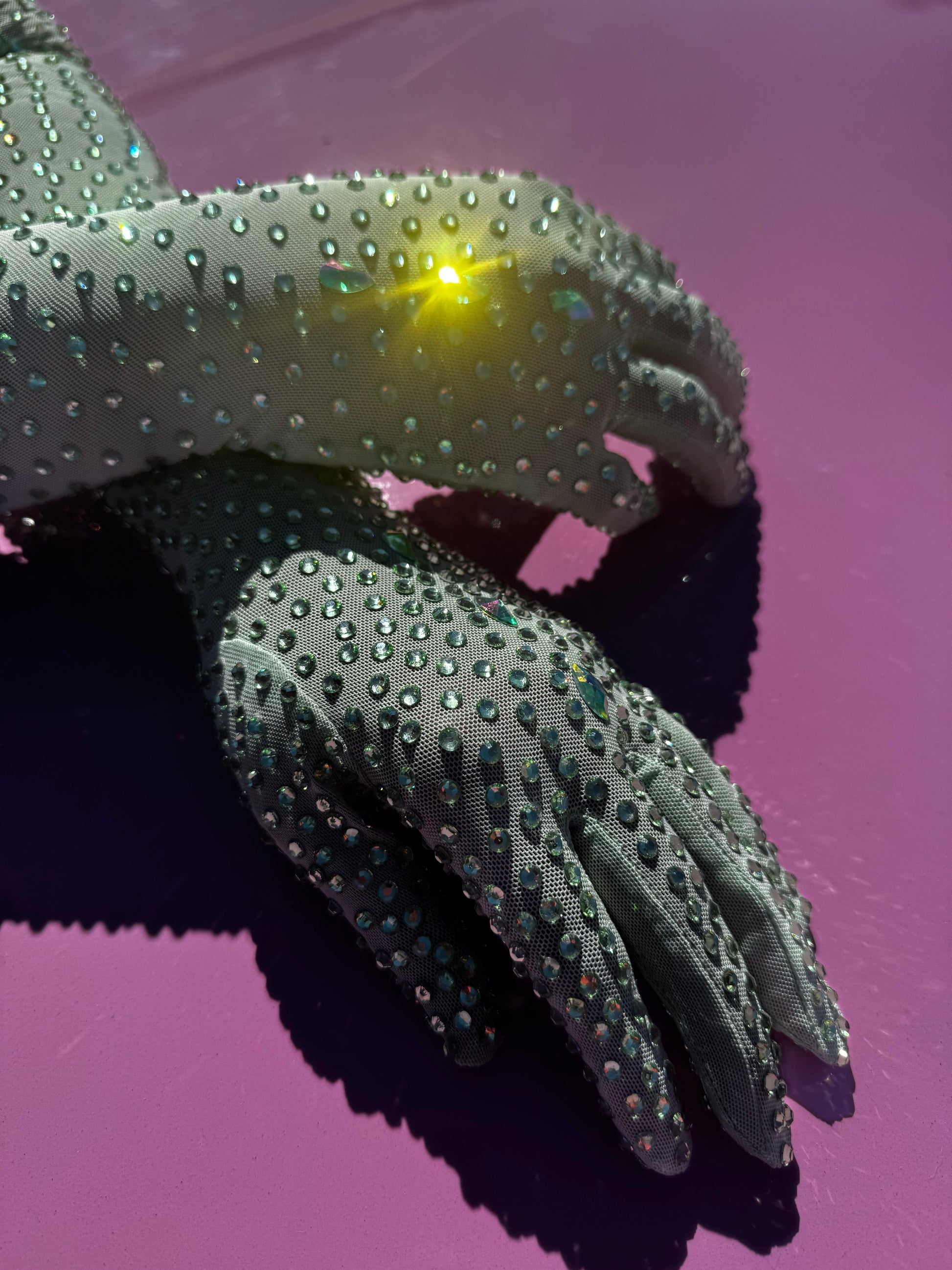 A pair of hands wearing Viva La Bombshell Burlesque's The Mint Special - Rhinestone Gloves rest on a glossy, bright purple surface, while a small light reflects off one opera-length glove, creating a striking starburst.