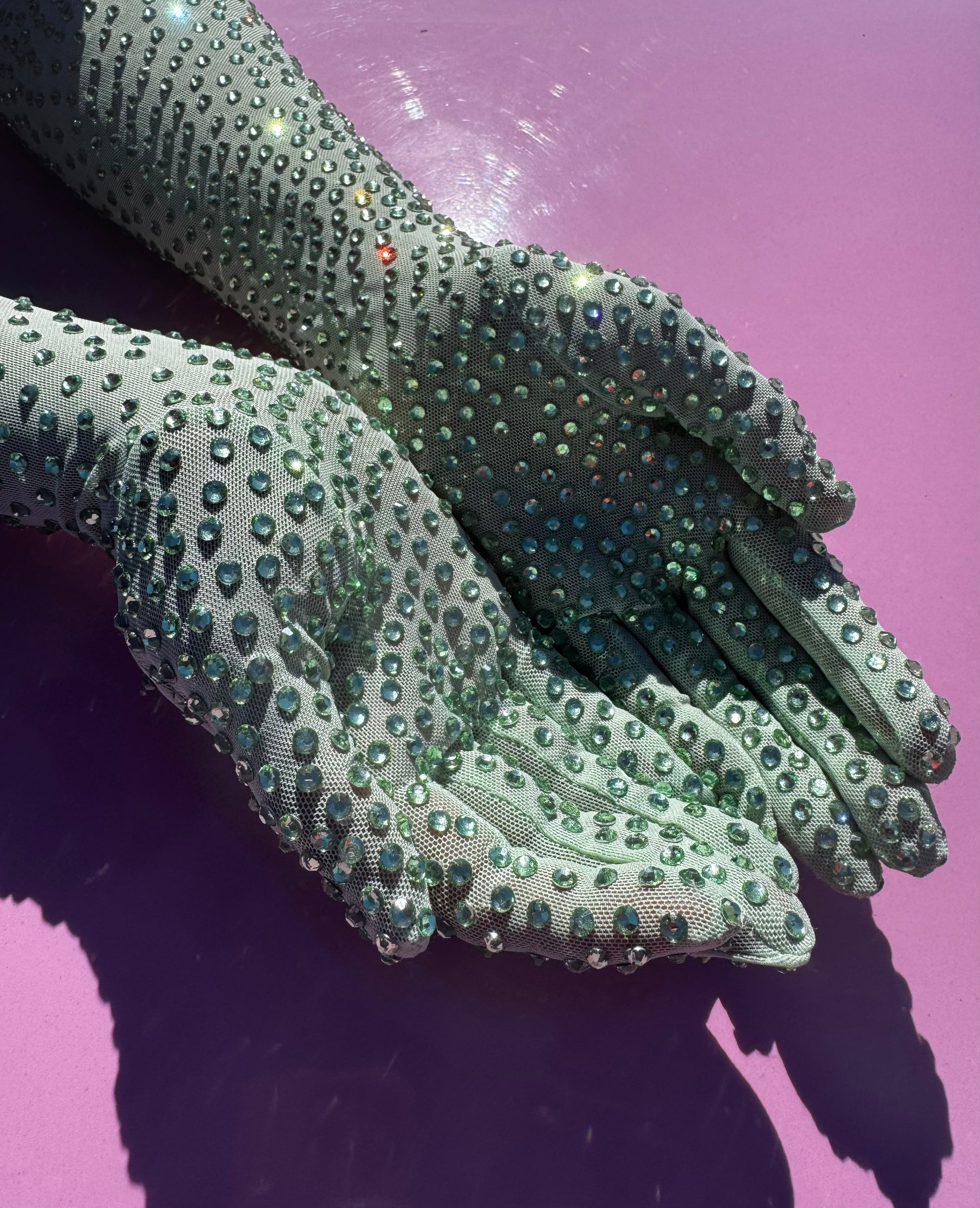 Two hands wearing Viva La Bombshell Burlesque’s The Mint Special - Rhinestone Gloves are held together on a shiny lavender surface, reflecting light and creating a shimmering effect.