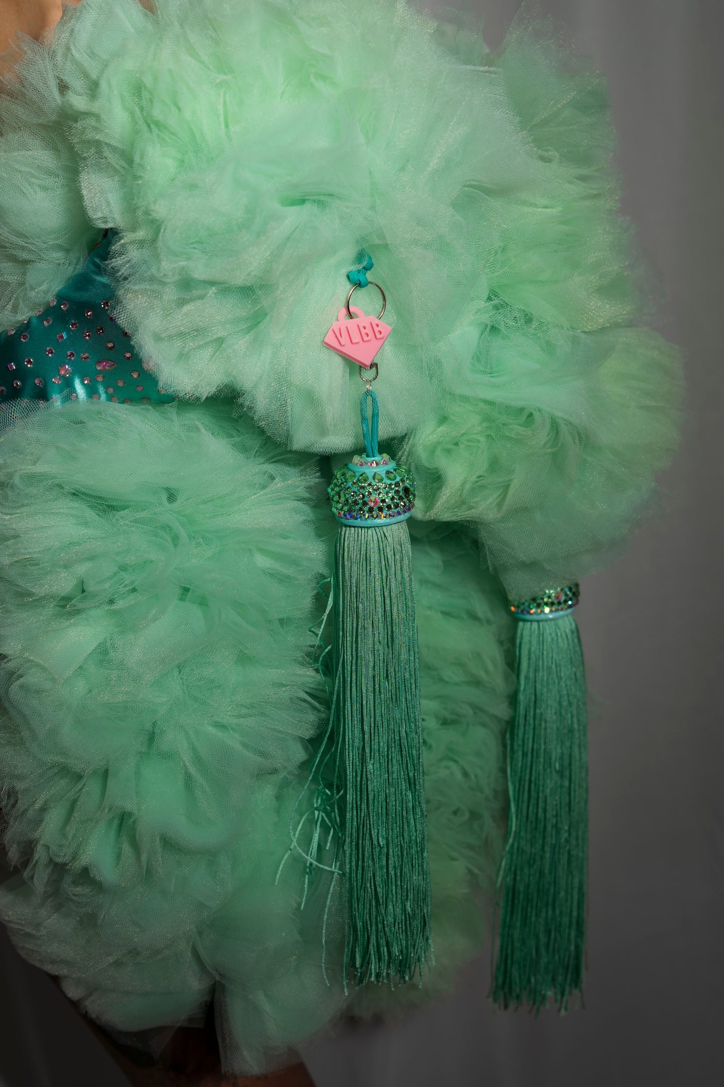 A close-up of The Dazzle - Tulle Boa by Viva La Bombshell Burlesque, featuring vibrant mint green tulle and two long matching tassel toppers adorned with rhinestones and a pink VLED charm, hanging elegantly from the made-to-order fabric.
