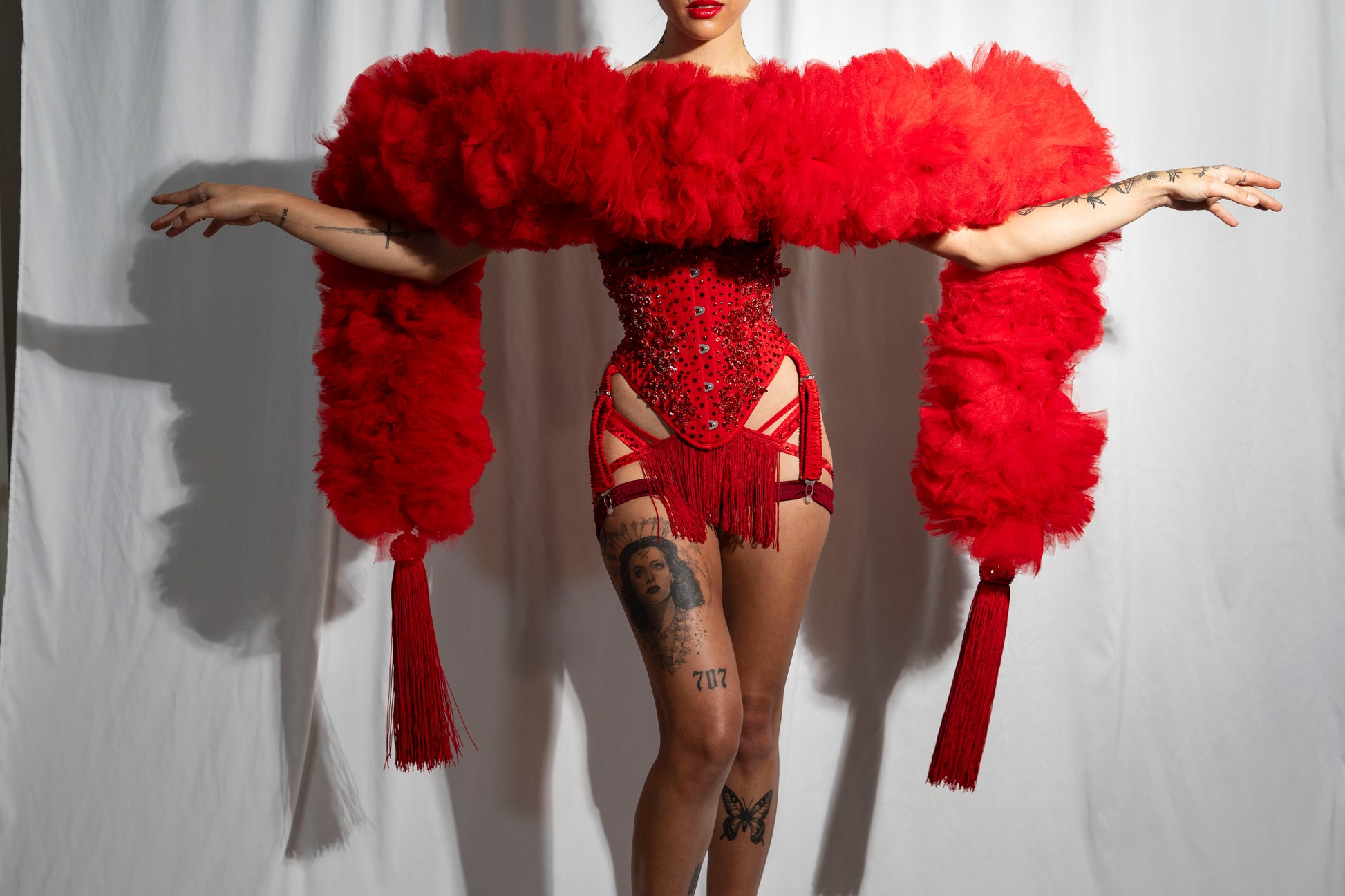 Person poses with arms outstretched, wearing Viva La Bombshell Burlesque's The Dazzle - Tulle Boa. Bold red ruffles, tassel toppers, and sequins shimmer over tattooed skin. A soft white drape sets the background. This striking look is made to order.