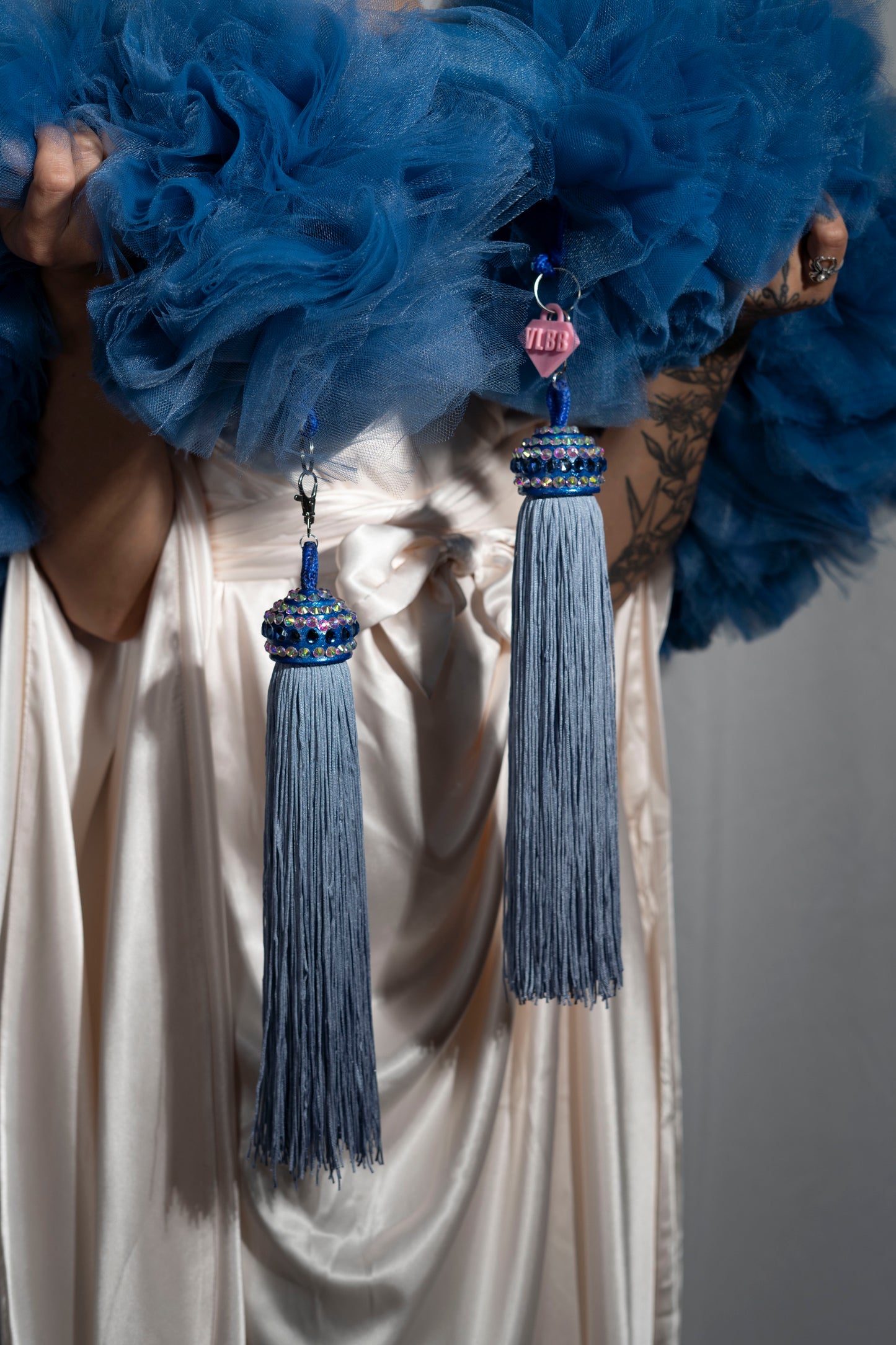 A tattooed person in a cream satin dress holds Viva La Bombshell Burlesque’s The Dazzle – Tulle Boa, featuring blue tulle, two long blue beaded tassel toppers, and a pink acrylic VIBE heart charm. Each piece is made to order.