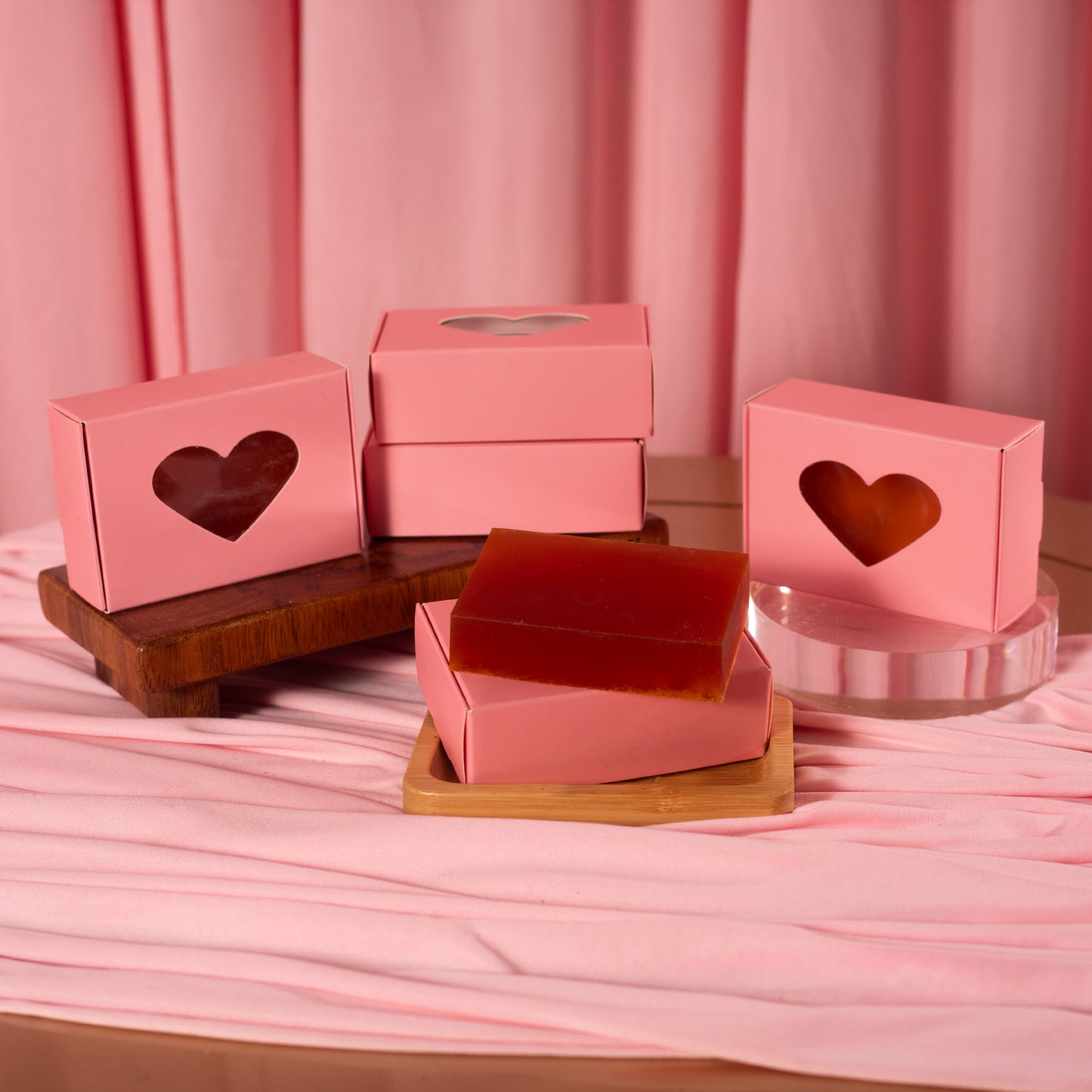 Several pink boxes with heart cutouts are shown on wooden and clear stands, surrounding Viva La Bombshell Burlesque Turmeric Soap bars for moisturized skin, all set against a pink fabric backdrop with matching curtains.