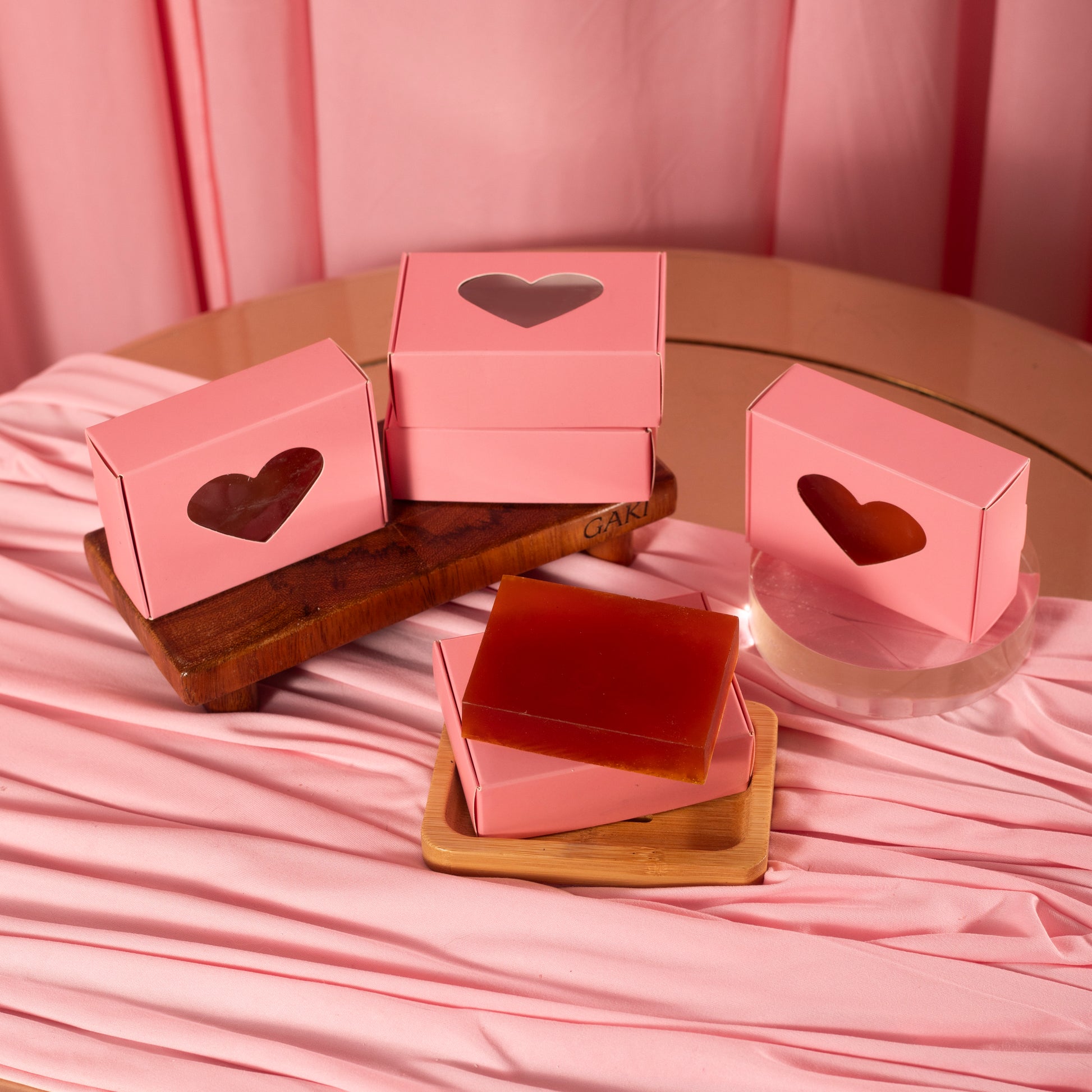 Viva La Bombshell Burlesque Turmeric Soap, in pink rectangular boxes with heart-shaped windows, is displayed on a draped pink surface. Some boxes are open, showing the reddish-orange soap for even tone and moisturized skin. Pink curtain behind.