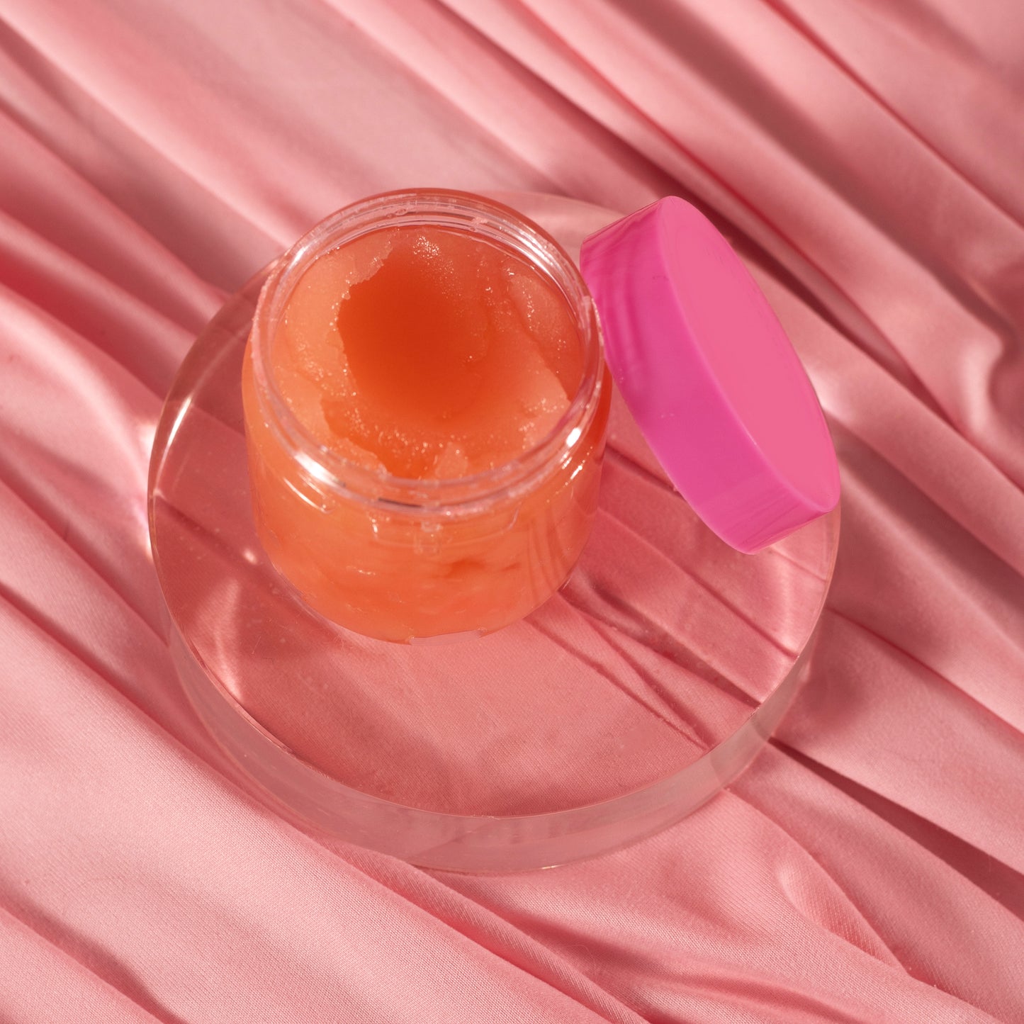 A small open jar of Viva La Bombshell Burlesque Diamond Lip Scrub with a pink lid sits on a clear round platform atop soft, pink satin-like fabric.