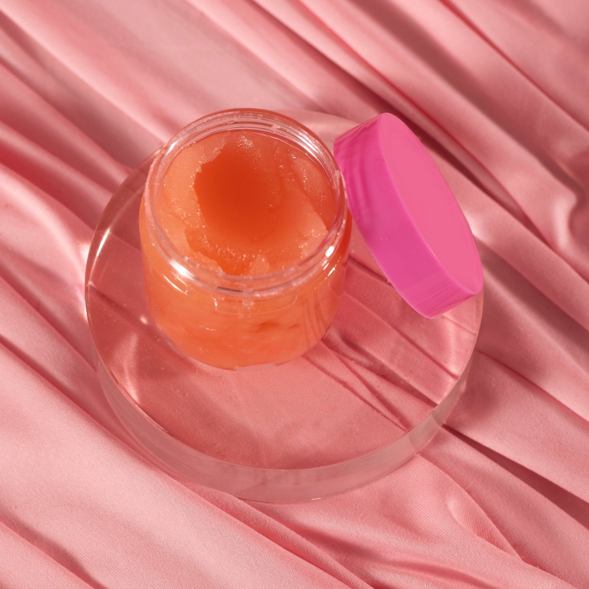 A small open jar of Viva La Bombshell Burlesque Diamond Lip Scrub with a pink lid sits on a clear round platform atop soft, pink satin-like fabric.