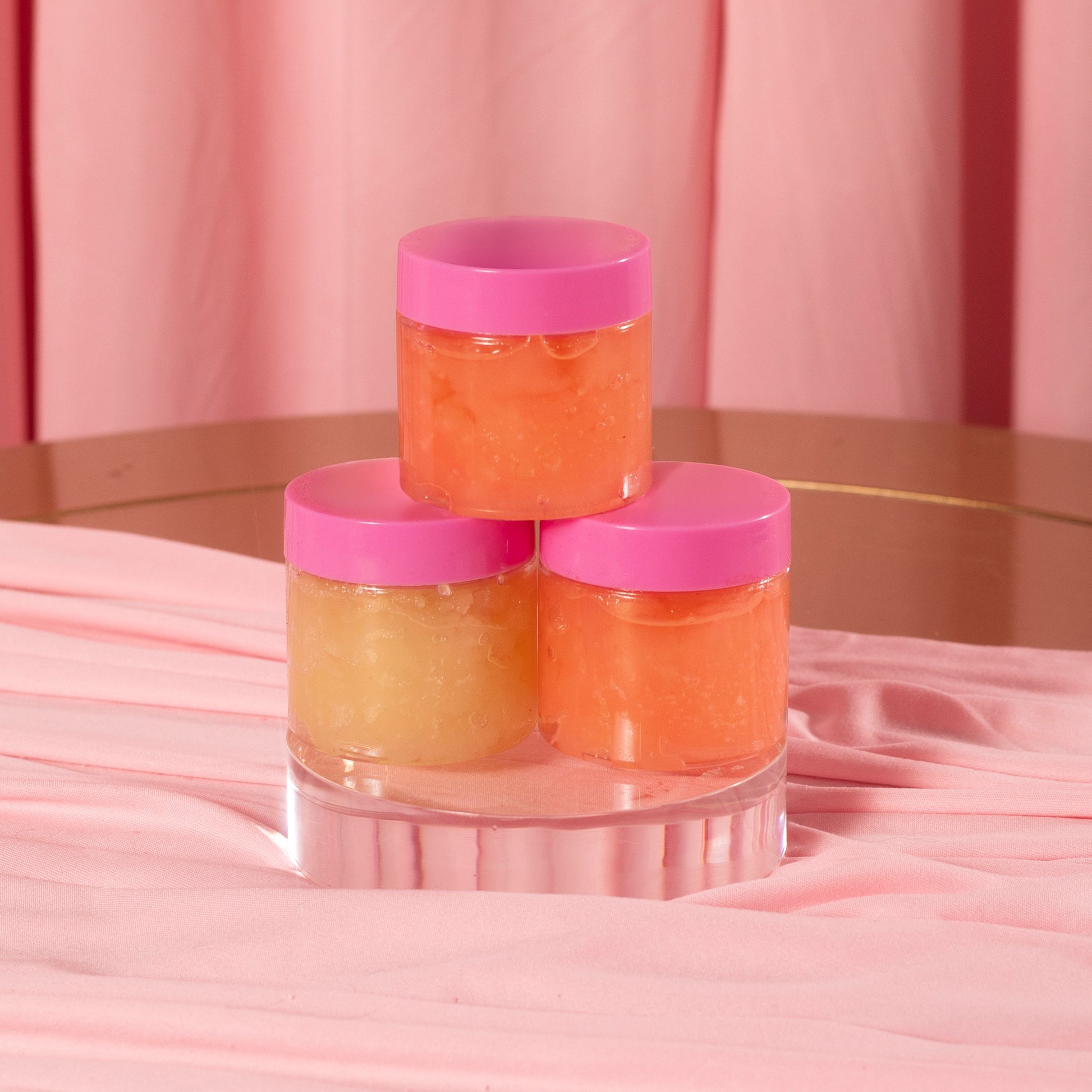 Three small jars of Viva La Bombshell Burlesque's Diamond Lip Scrub, each with a pink lid, rest on a clear pedestal. Their yellow and orange hues stand out against the soft pink background and fabric for a coordinated display.