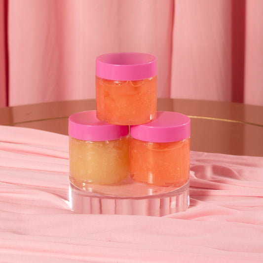 Three small jars of Viva La Bombshell Burlesque's Diamond Lip Scrub, each with a pink lid, rest on a clear pedestal. Their yellow and orange hues stand out against the soft pink background and fabric for a coordinated display.