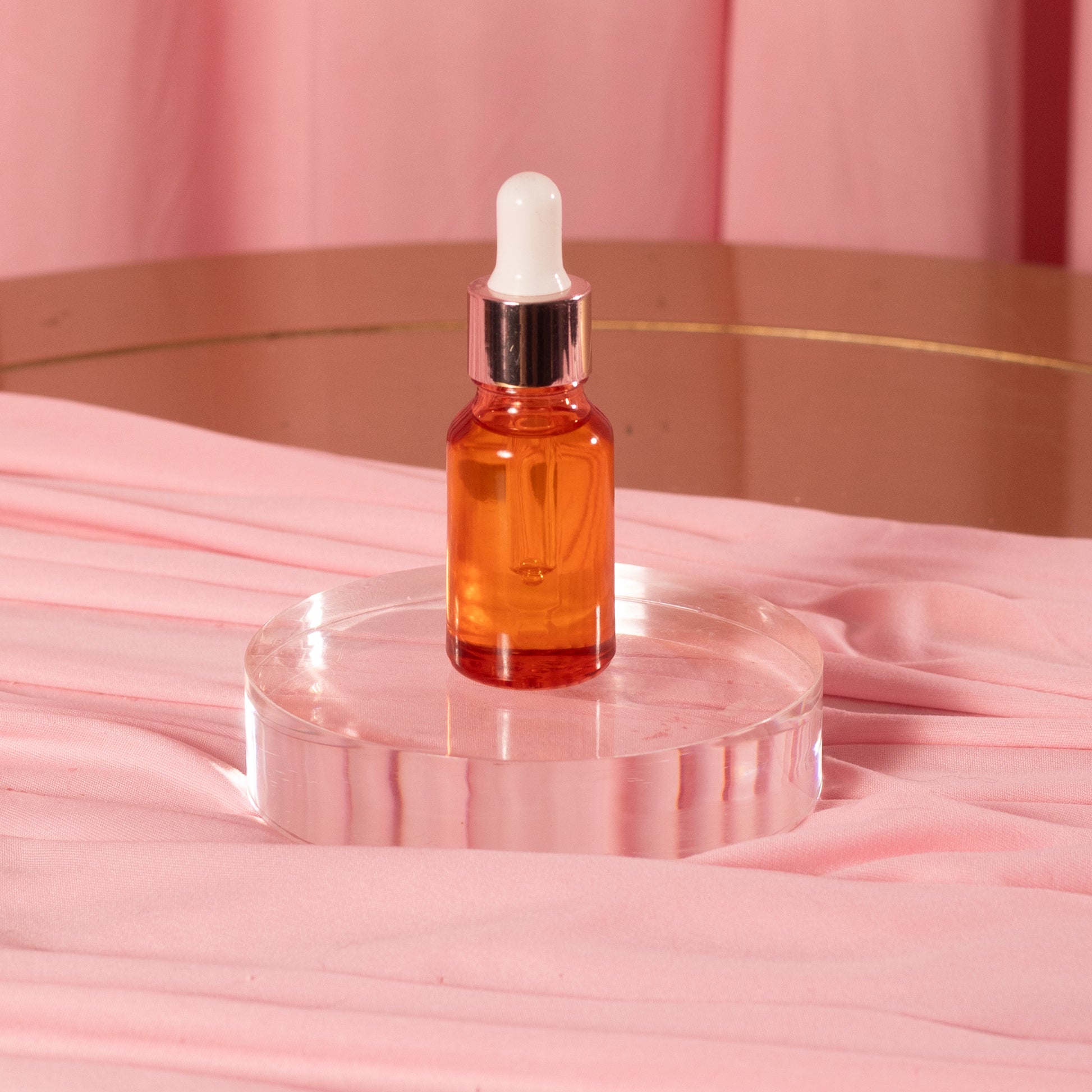 A small amber glass dropper bottle of Viva La Bombshell Burlesque Cuticle Oil with a white cap sits on a clear round stand atop pink fabric, set against a backdrop of pink curtains.