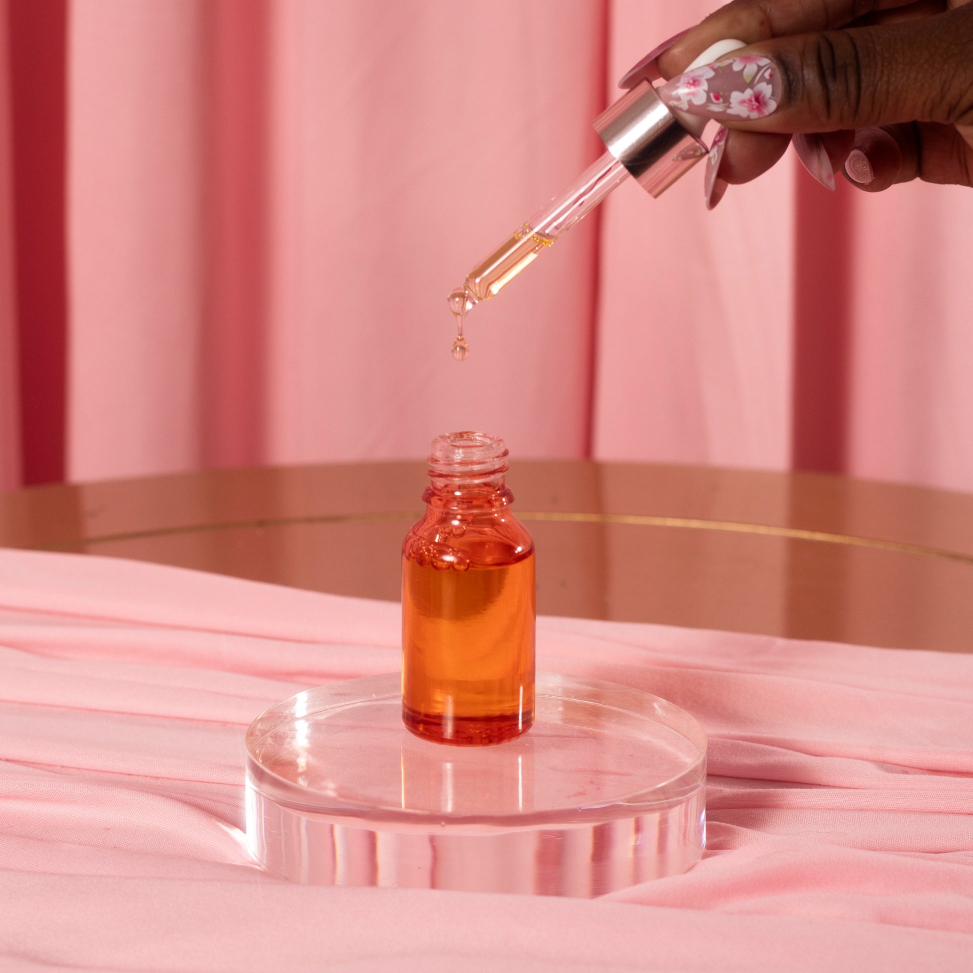 A hand with long, decorated nails holds a Viva La Bombshell Burlesque Cuticle Oil dropper above its small glass bottle of orange liquid on a clear circular stand, set against a pink fabric background.