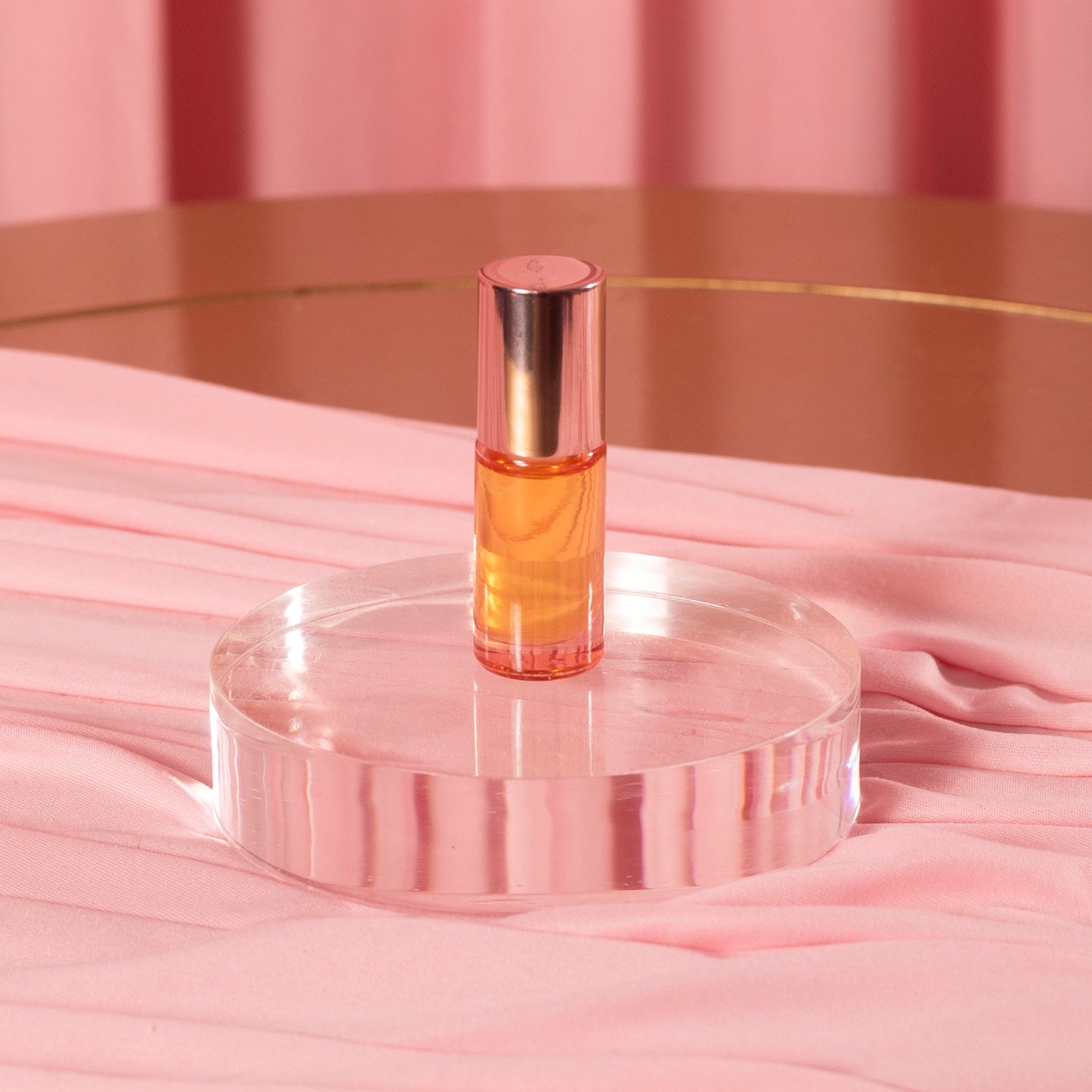 A small glass bottle of Viva La Bombshell Burlesque Cuticle Oil with a rose gold cap and orange Manuka oil sits on a clear round platform, set against pink fabric on a gold-rimmed table.