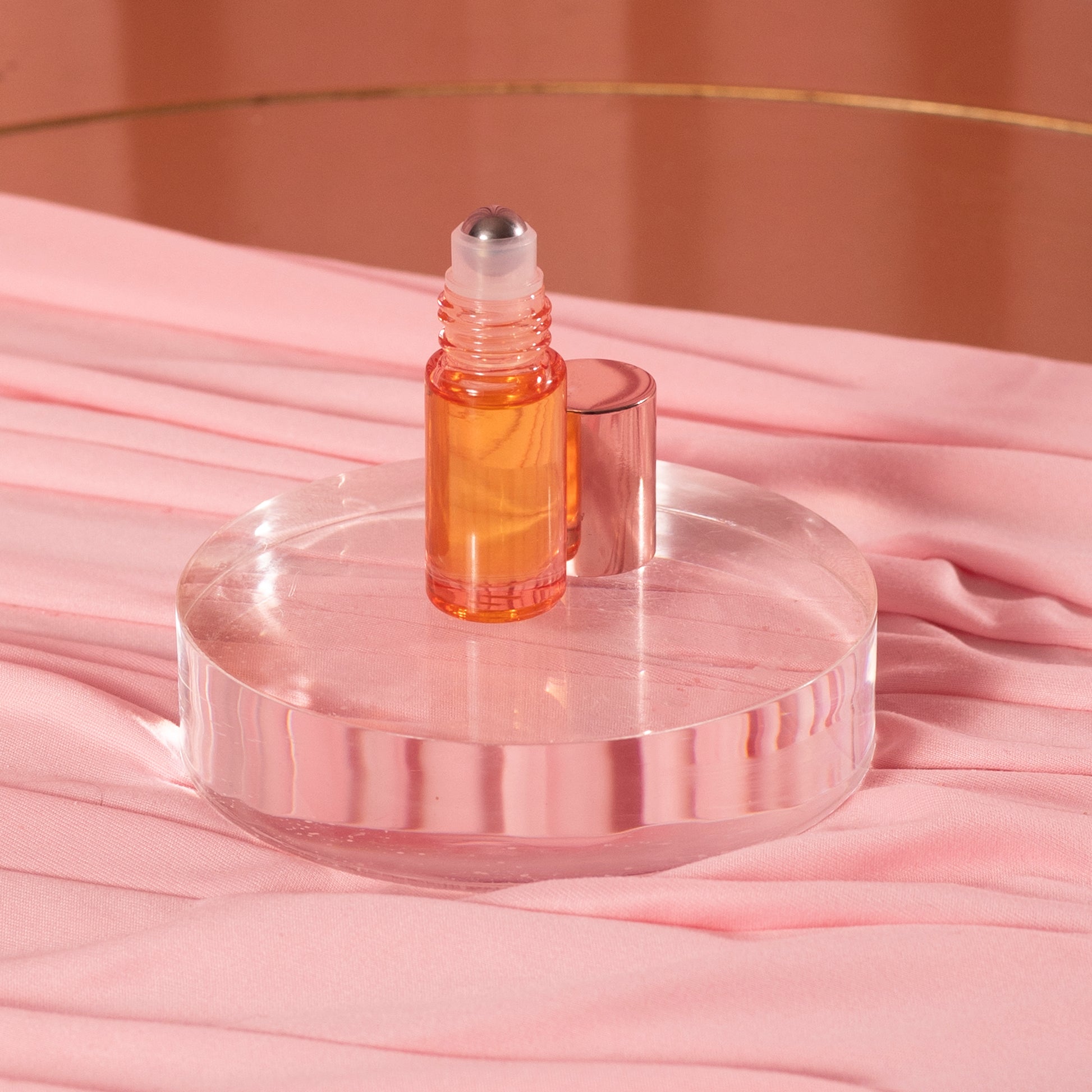 A small Viva La Bombshell Burlesque Cuticle Oil bottle with orange liquid stands on a clear round platform, its rose gold cap nearby. The setup is displayed against a pink, silky fabric background.