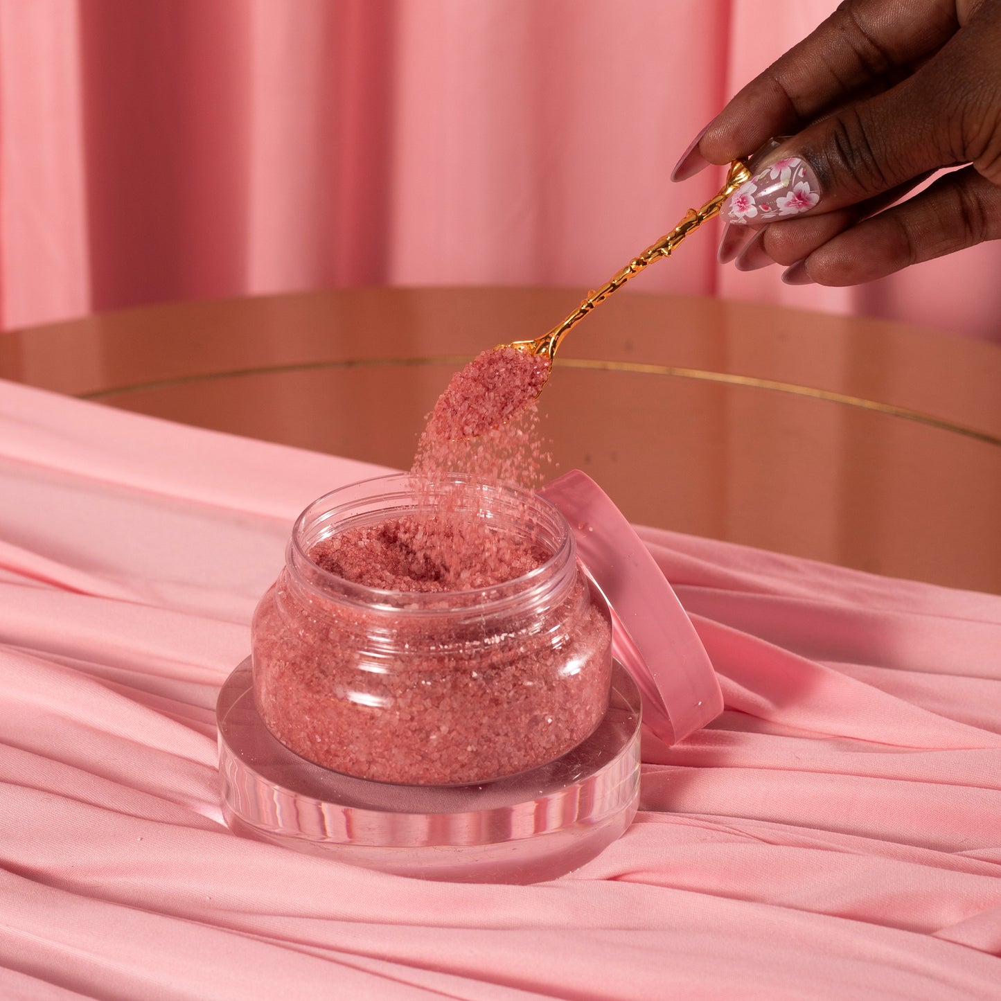 A manicured hand holds a gold spoon, scooping Viva La Bombshell Burlesque’s Diamond Salt Soak—pink scrub with Dead Sea salt—from a clear jar on pink fabric, set against a pink curtain backdrop.
