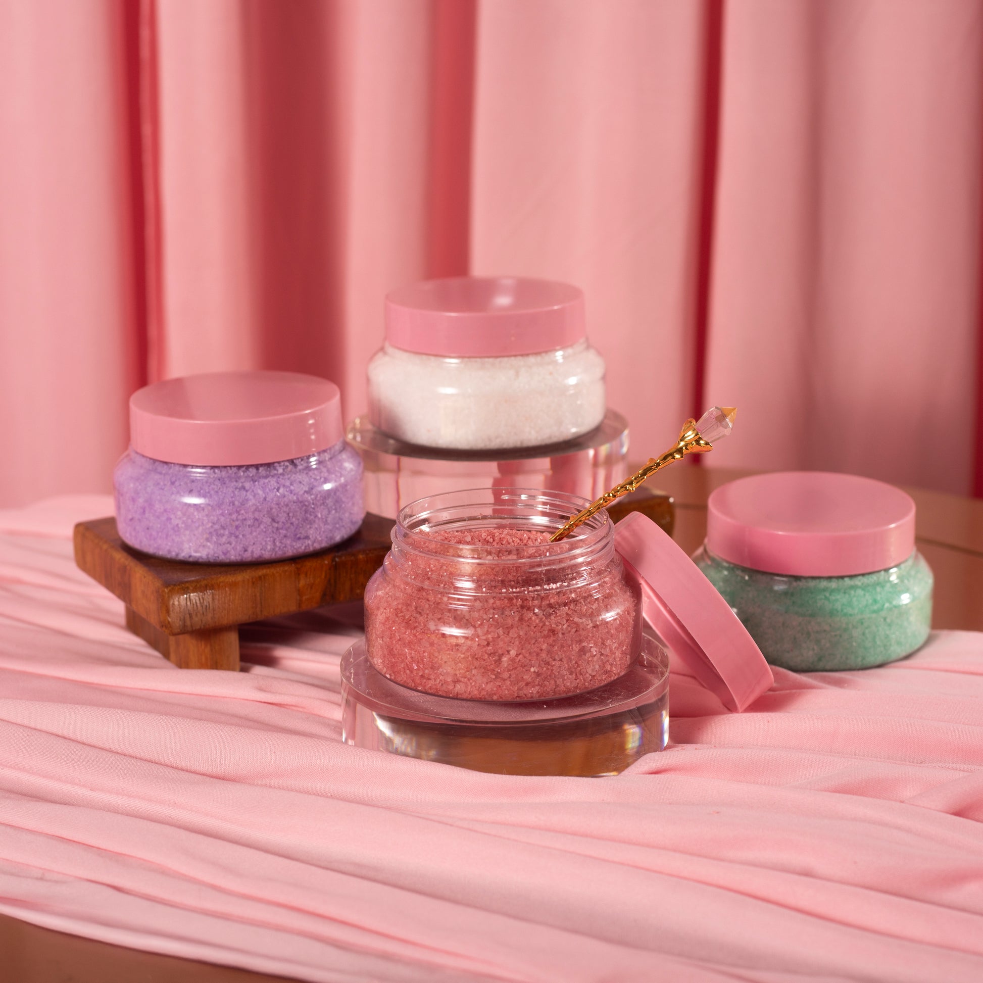 Four jars of Viva La Bombshell Burlesque Diamond Salt Soak in pink, purple, white, and green with pink lids rest on pink fabric. One jar of pink salt is open with a gold spoon. A wooden stand holds two jars behind.