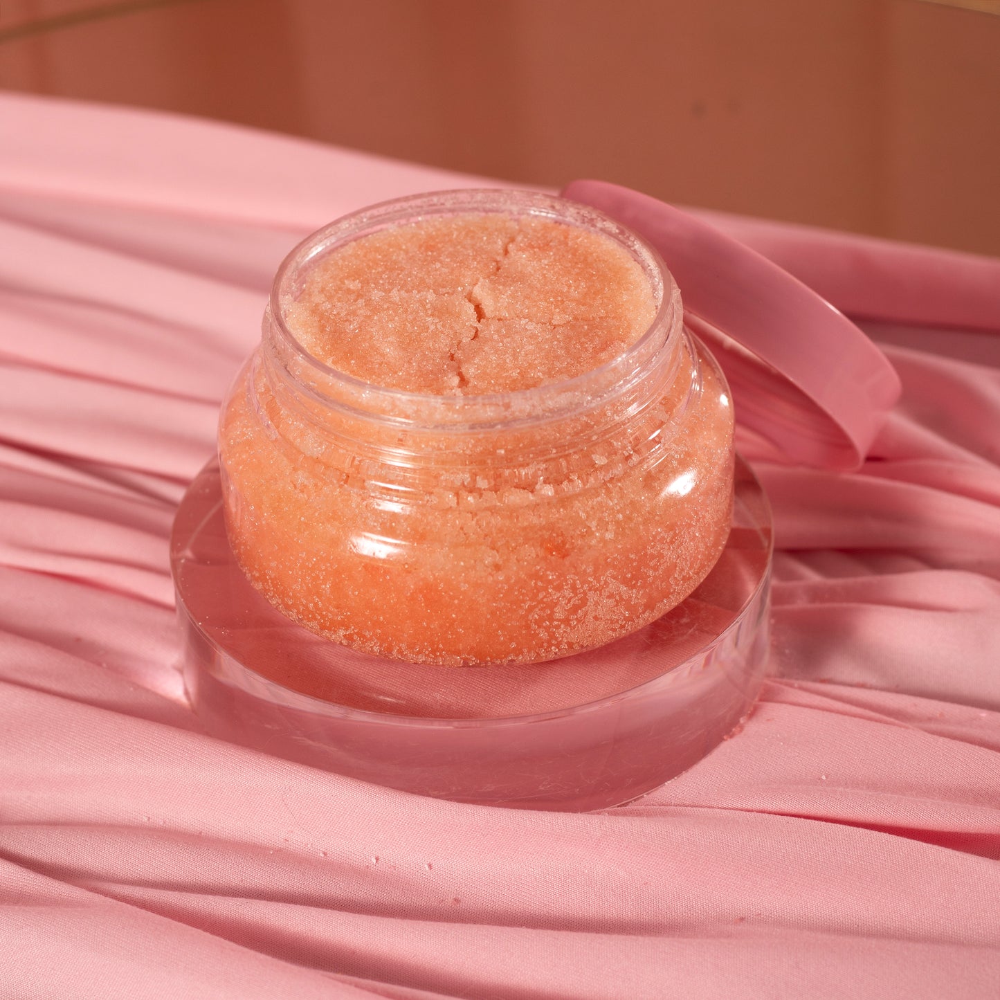 An open jar of Viva La Bombshell Burlesque Diamond Body Scrub, peach-colored with a pink lid, rests on a round acrylic tray atop soft pink fabric.