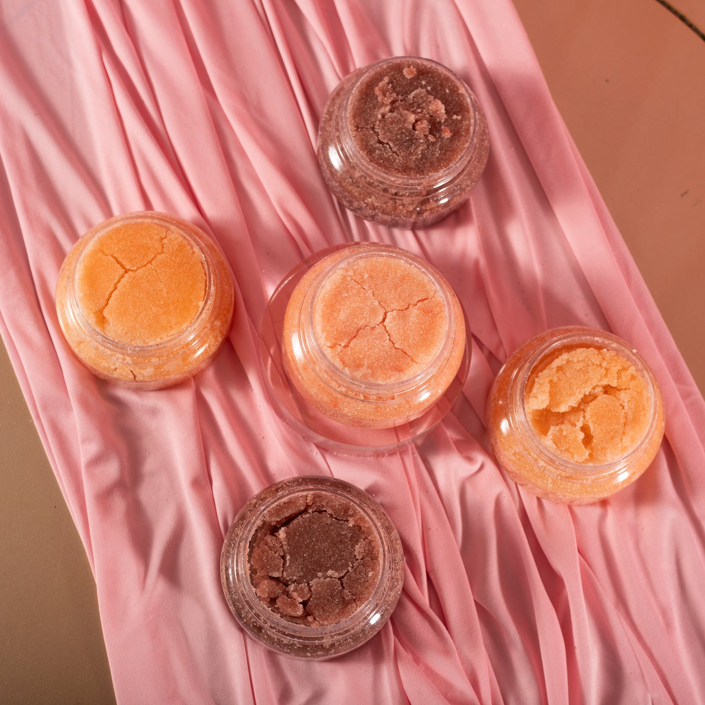 Five jars of Viva La Bombshell Burlesque's Diamond Body Scrub in orange and brown tones sit on pink fabric, creating a soft, textured backdrop that highlights the round jars.