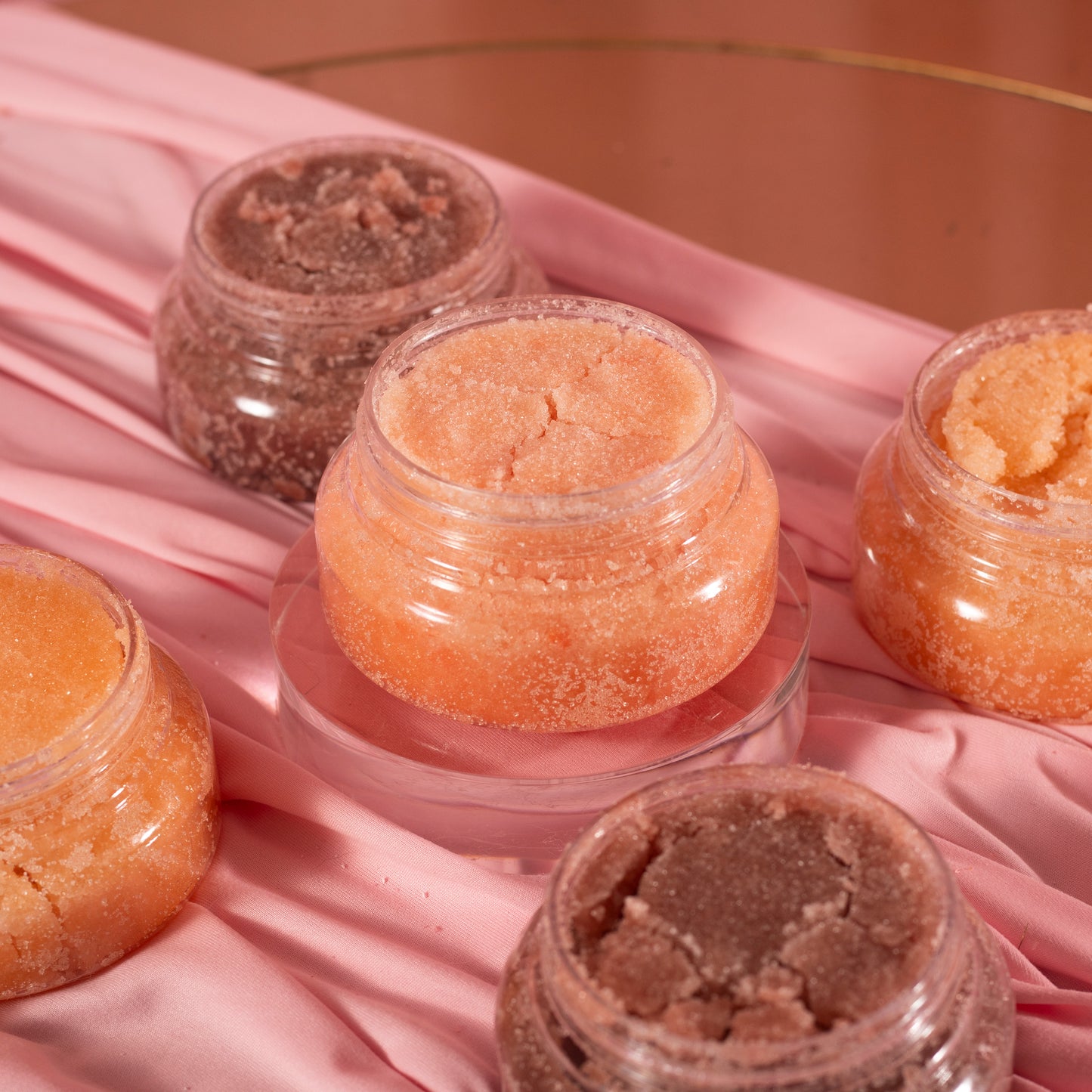 Five jars of Viva La Bombshell Burlesque’s Diamond Body Scrub in peach and brown tones are arranged on pink satin, with one elevated on a clear stand for a soft, luxurious display.