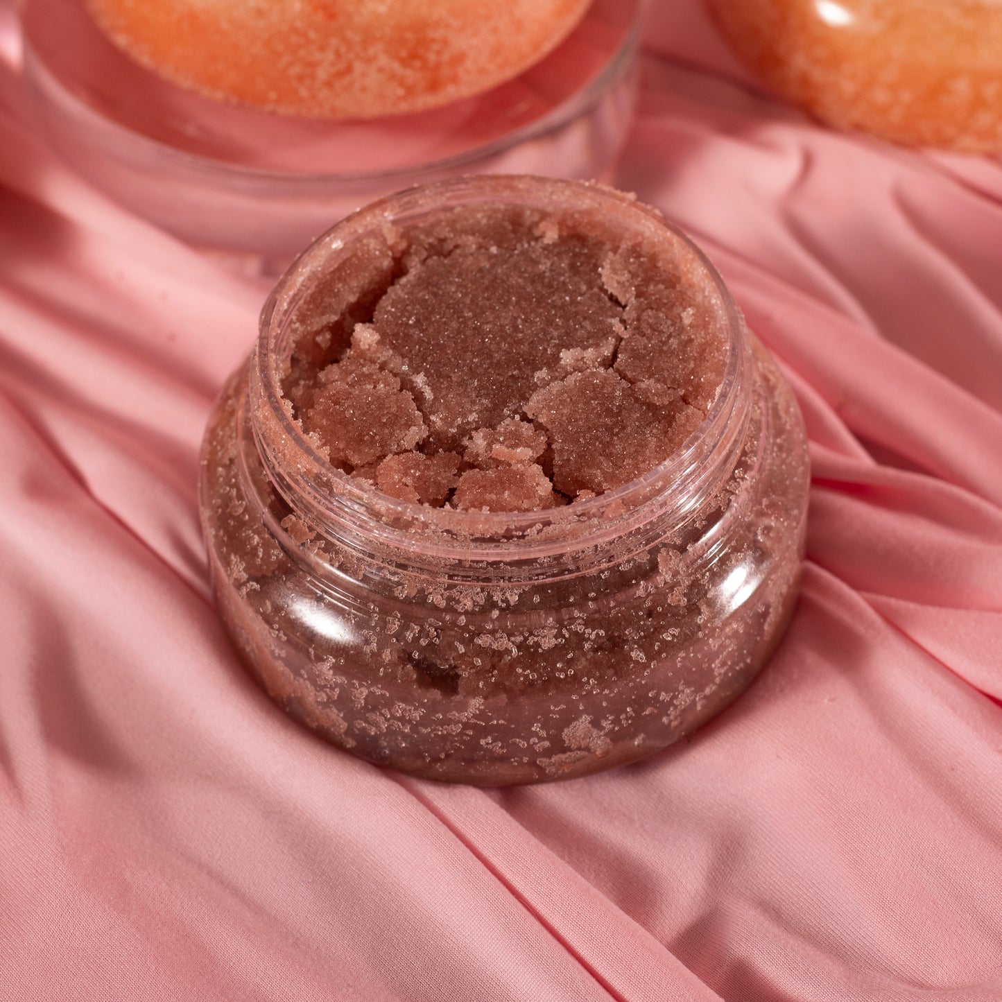 A clear jar of Viva La Bombshell Burlesque’s Diamond Body Scrub, a textured and grainy pink formula, rests on soft, wrinkled pink fabric.