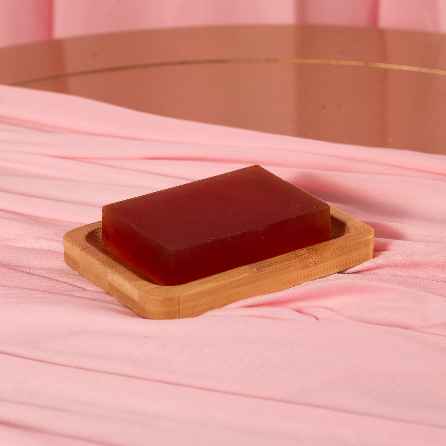 A rectangular red Viva La Bombshell Burlesque Turmeric Soap rests on a wooden dish atop pink fabric with a pink backdrop and round mirror—ideal for even-toned, moisturized skin.